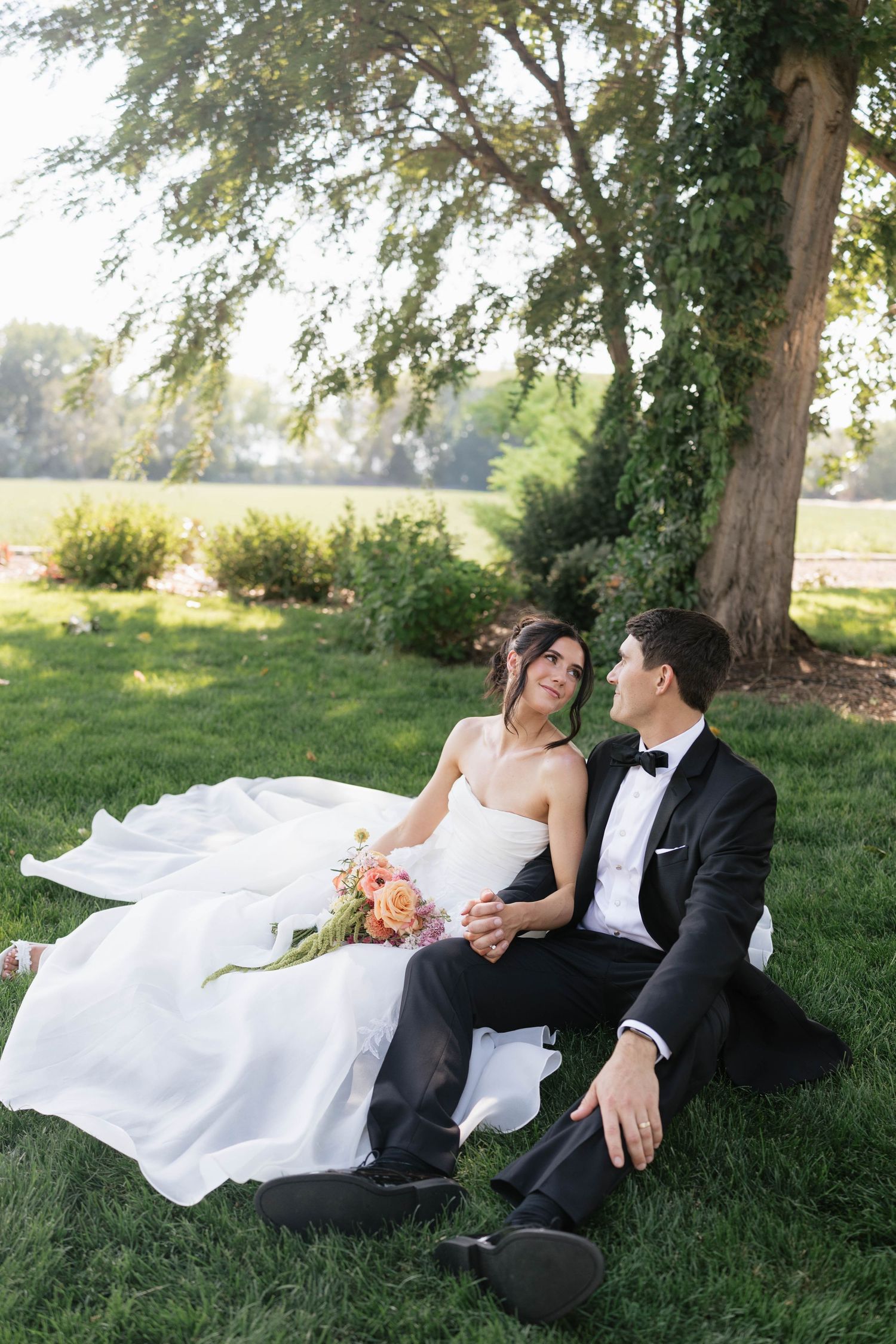 Bride and groom sitting on lush green grass under a large tree, holding hands and gazing at each other on their wedding day.