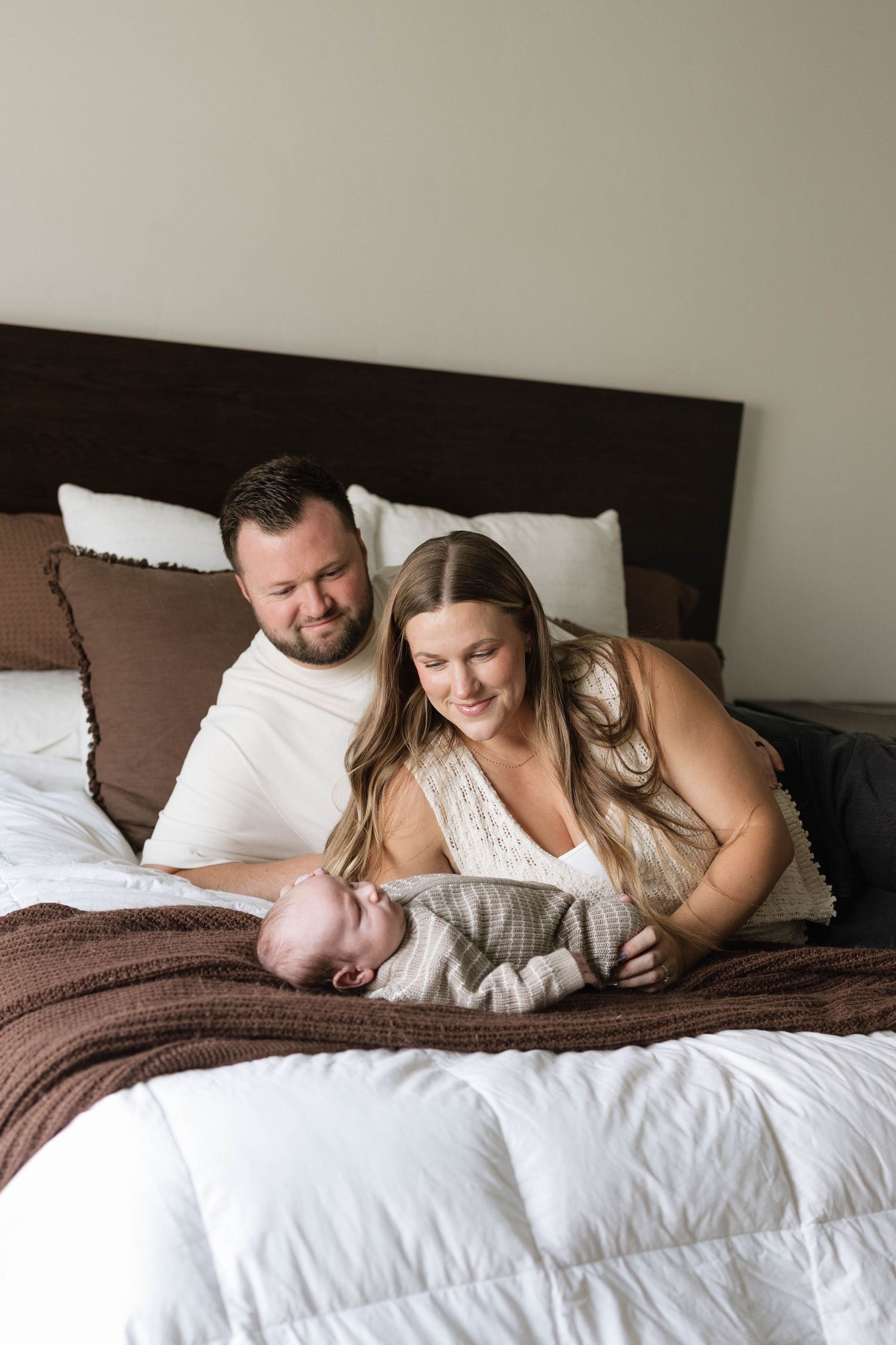 A couple smiles at their newborn baby while relaxing on a white bed with brown accents in a cozy bedroom.