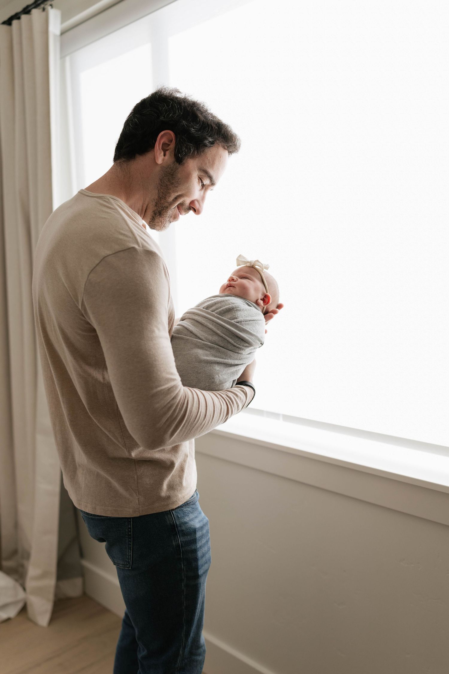 A father stands by a bright window, gently holding his swaddled newborn baby in his arms.