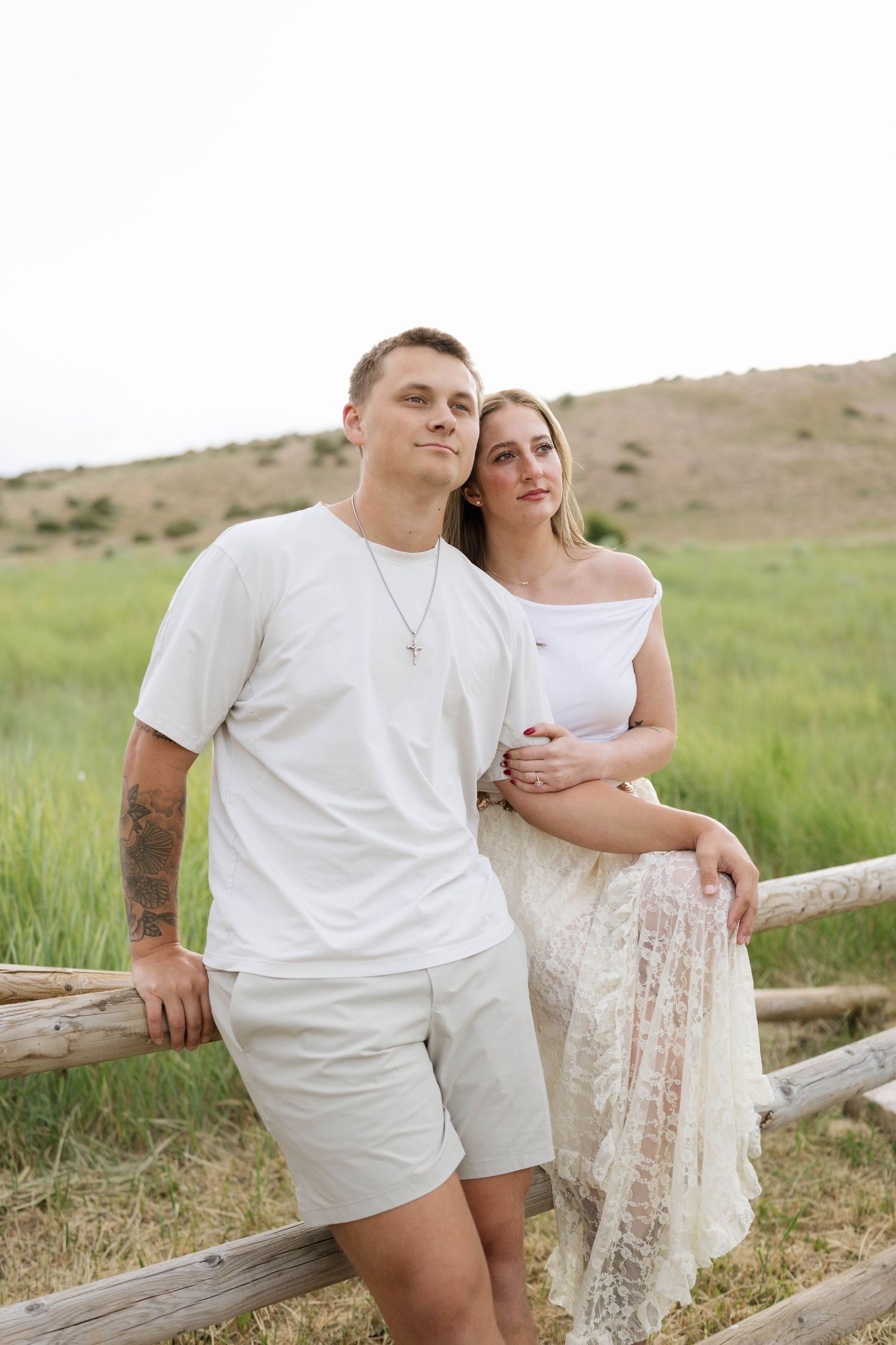 Couple in white outfits leaning on rustic wooden fence in green field, man with tattoo wearing cross necklace.