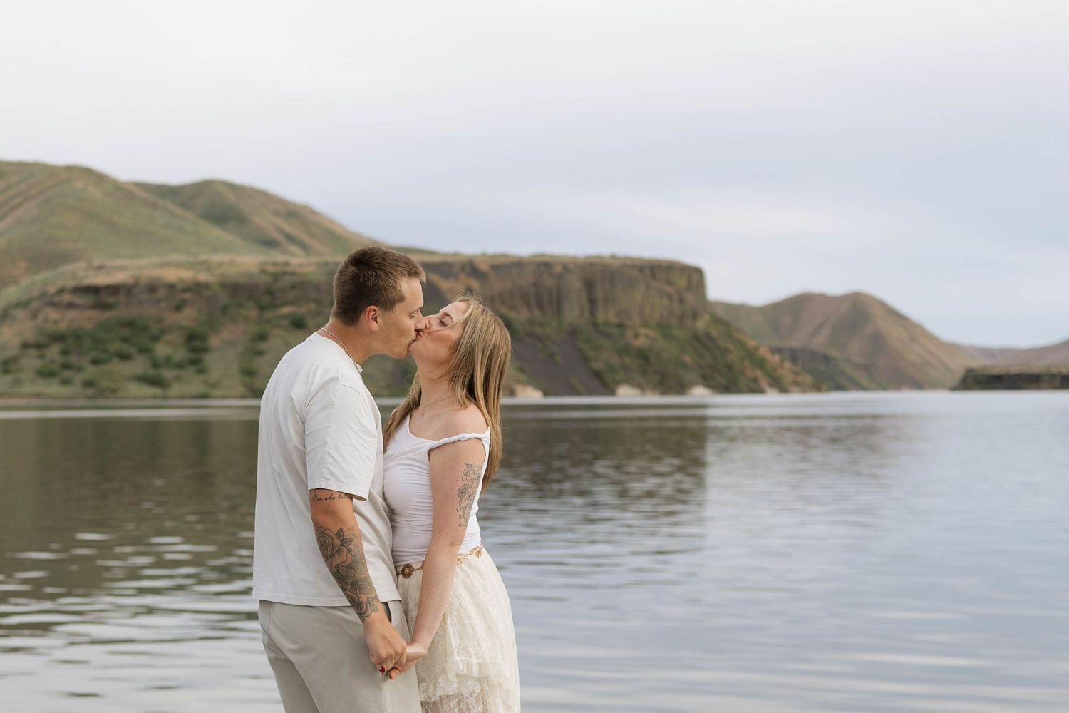 Couple sharing a romantic kiss by a serene lake with green hills in the background during an outdoor engagement session.