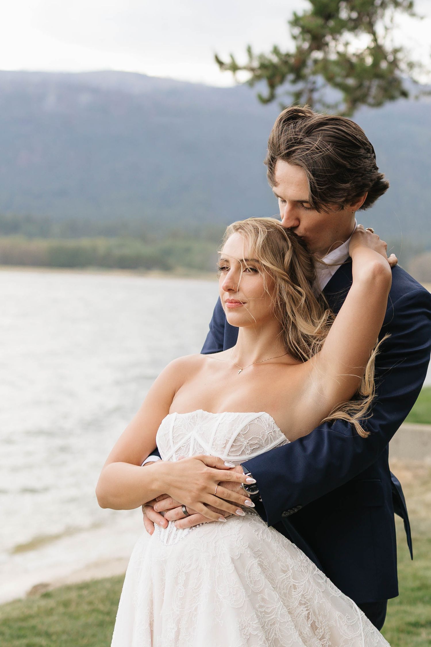 Groom in navy suit embraces bride in white lace gown by a mountain lake, sharing a tender kiss.