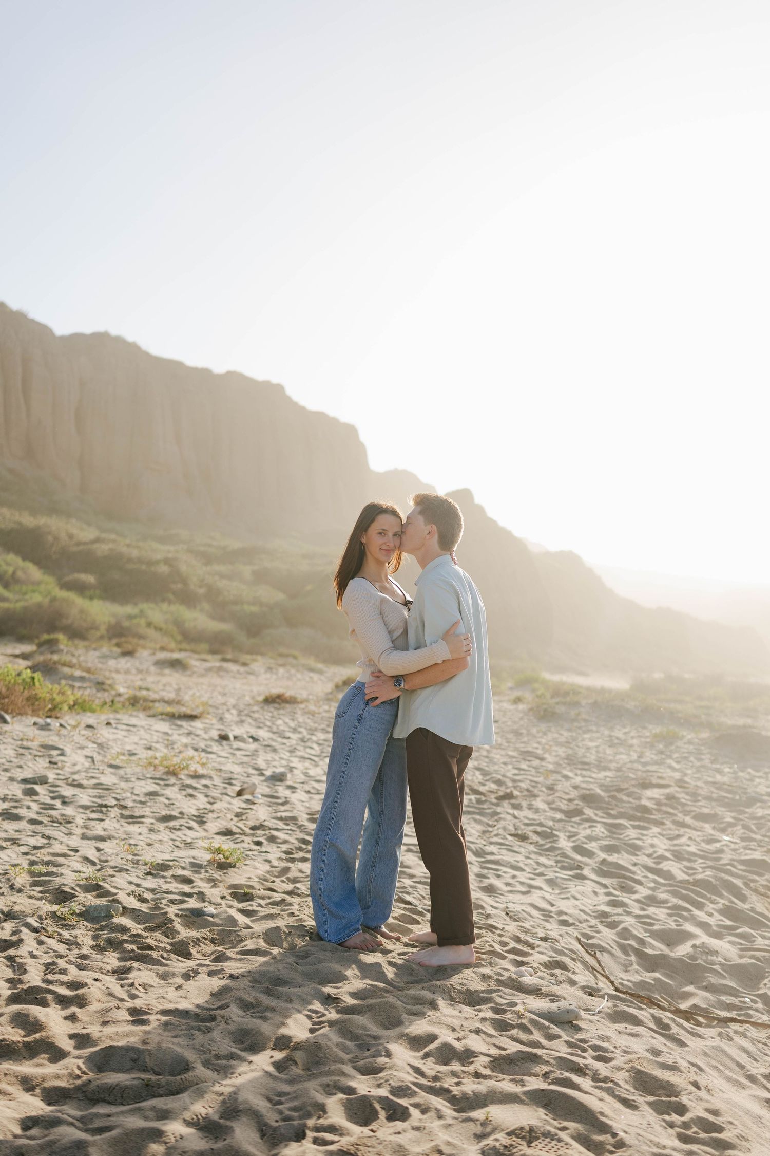 A couple shares a romantic moment on a sunlit beach with cliffs in the background during golden hour.