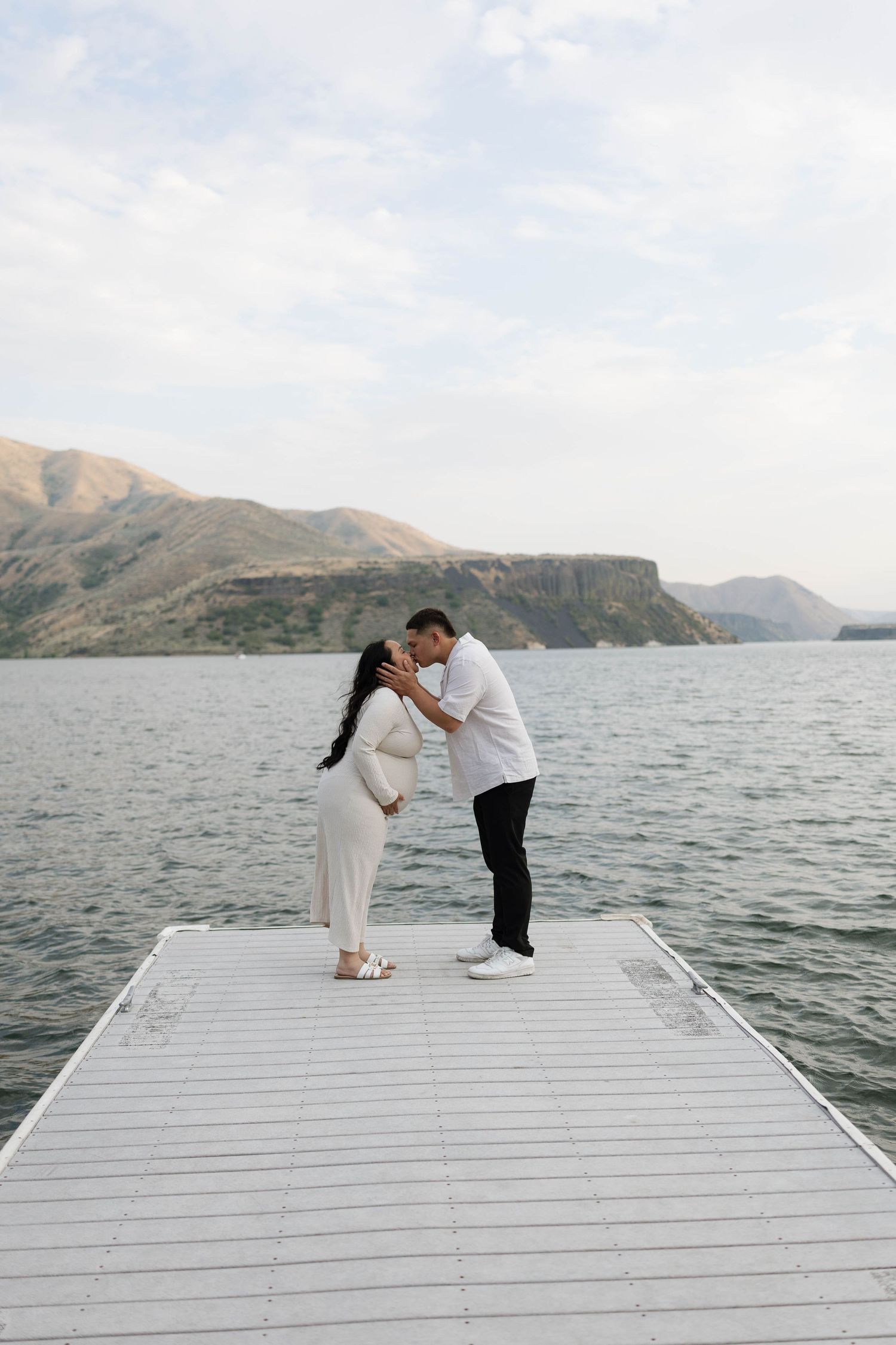 Pregnant couple sharing a kiss on a white dock overlooking a serene lake with mountain views.