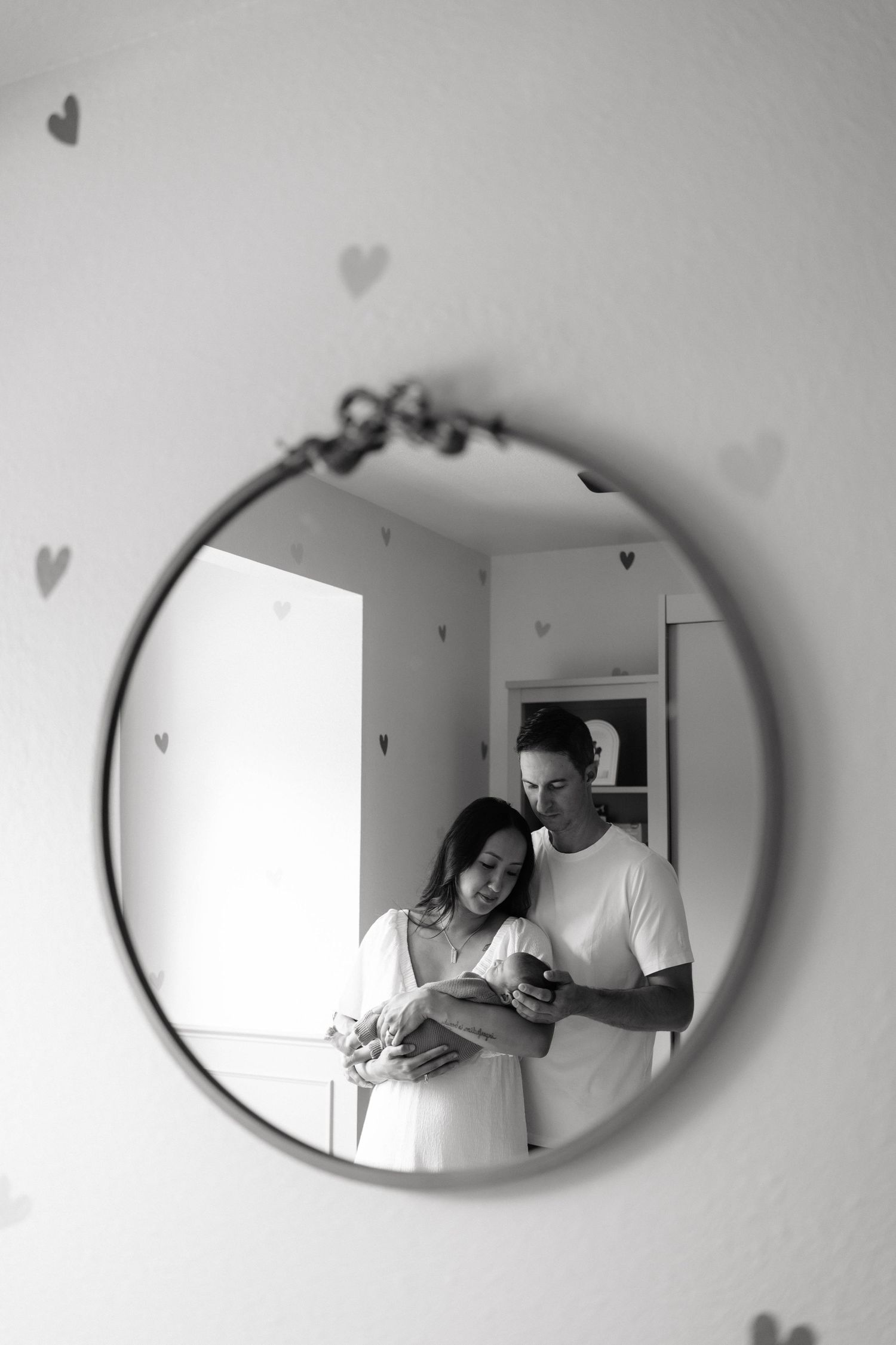 Black and white mirror reflection of parents cradling their newborn baby in a nursery with heart decals on the wall.