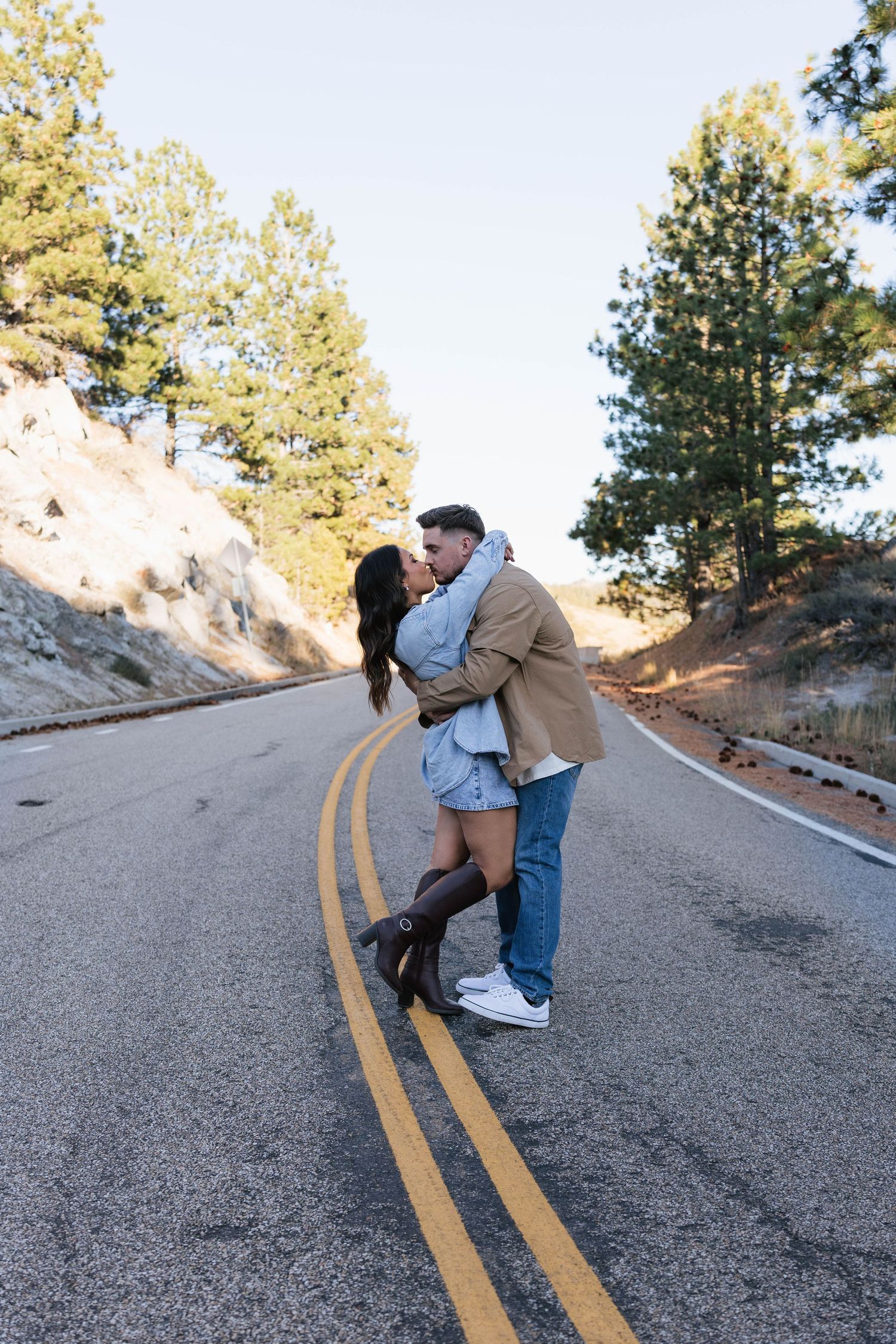A couple shares a romantic kiss on a winding mountain road surrounded by pine trees on a sunny day.