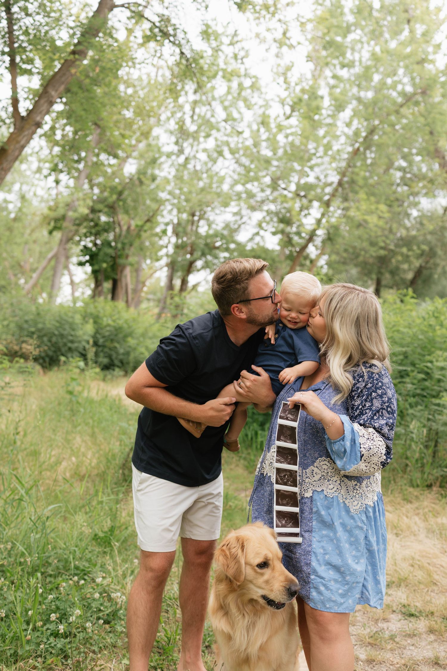 Family maternity photo with man kissing toddler, pregnant woman holding ultrasound, and golden retriever outdoors.