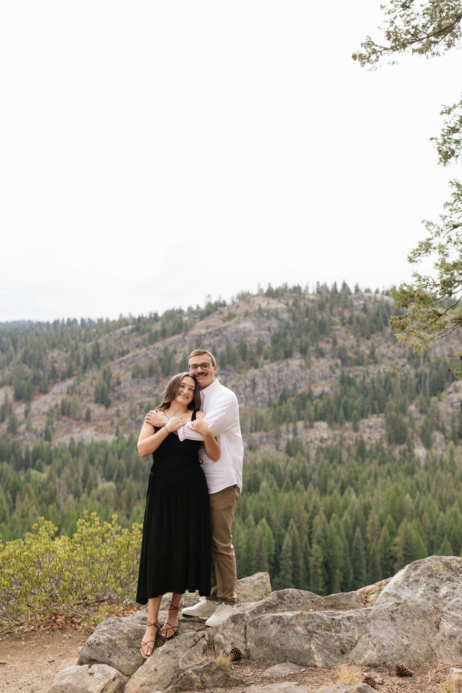 Couple embracing on a rocky overlook with a forested mountain valley behind them, a romantic engagement or outdoor portrait.