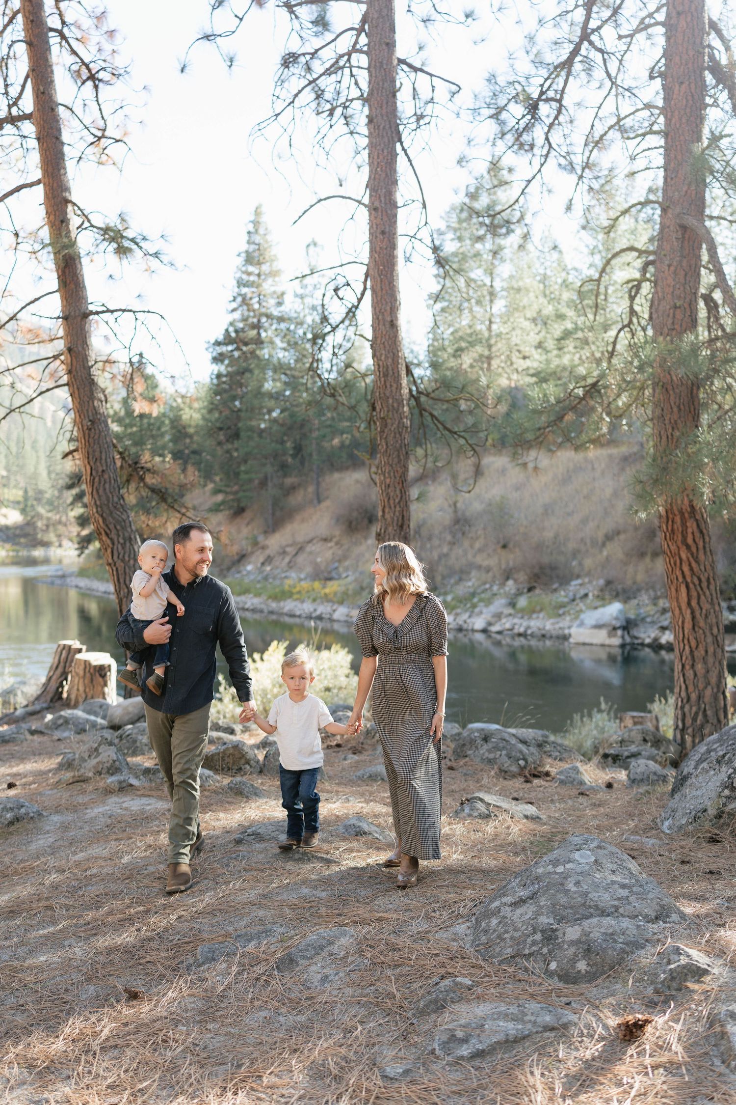 Family of four walking hand in hand along a rocky riverbank beneath tall pine trees during an outdoor family photo session.