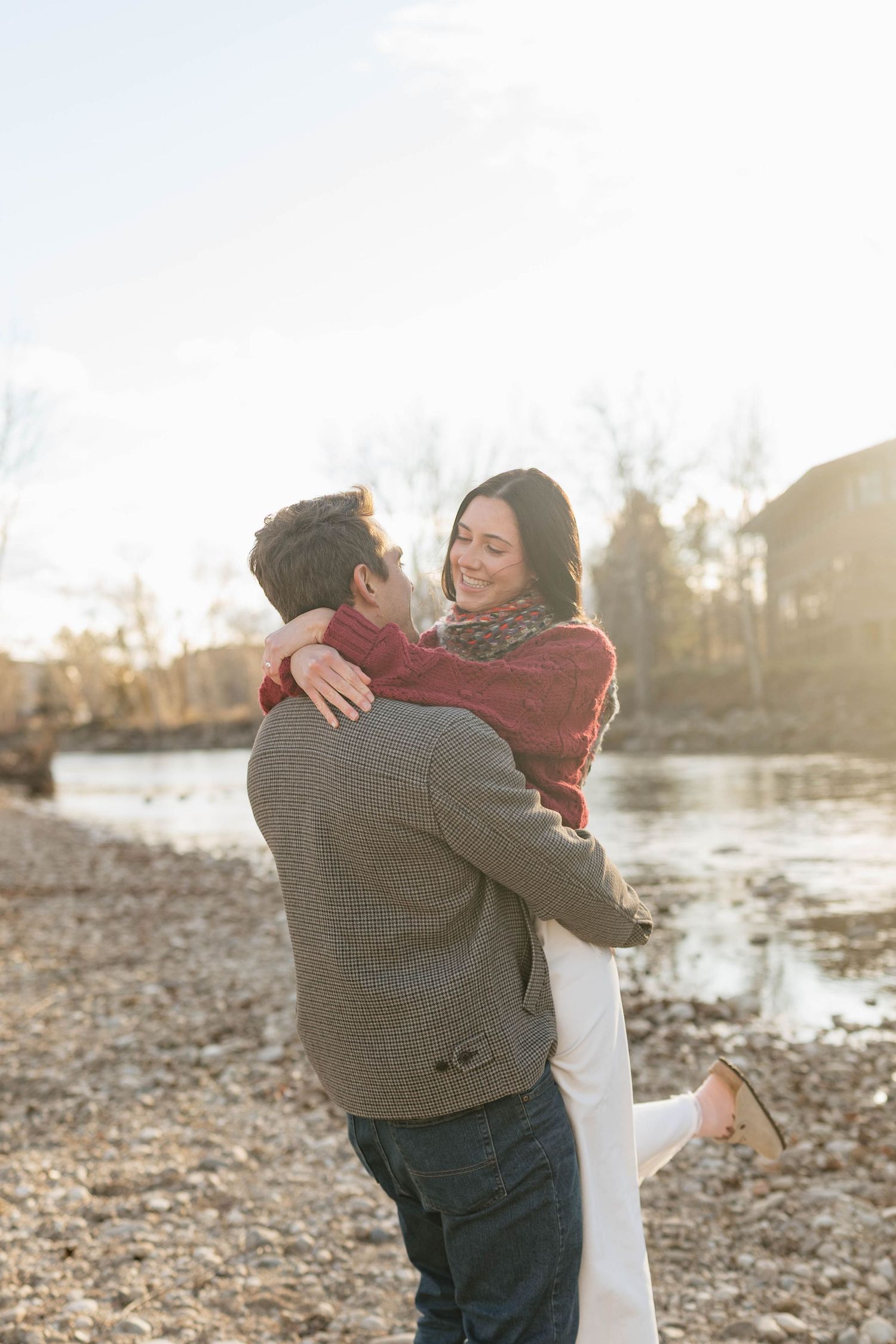 A couple embraces by a lake shore during golden hour with autumn leaves scattered on the ground.
