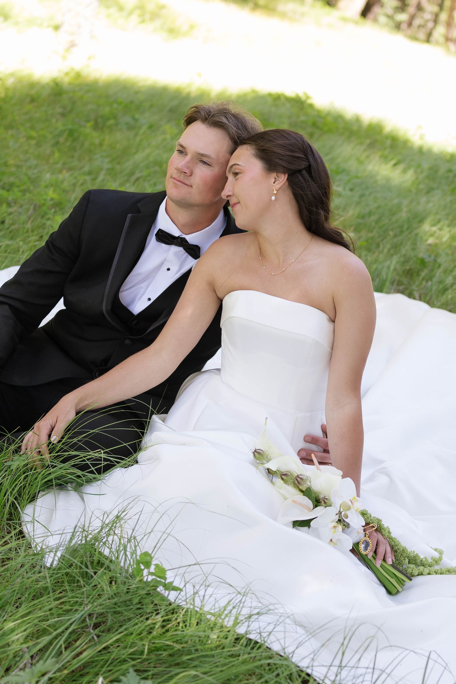 A bride and groom sitting together on green grass, wearing wedding attire, with the bride holding a white bouquet.