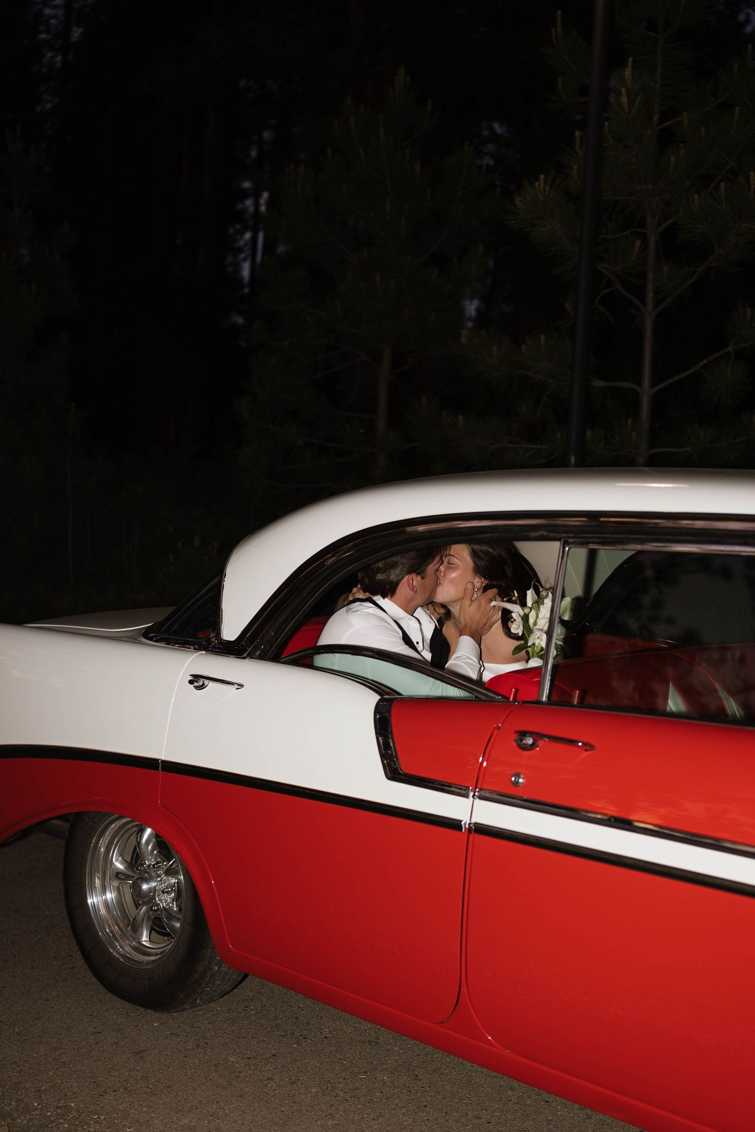 Newlywed couple sharing a romantic kiss inside a classic red and white vintage car at night.
