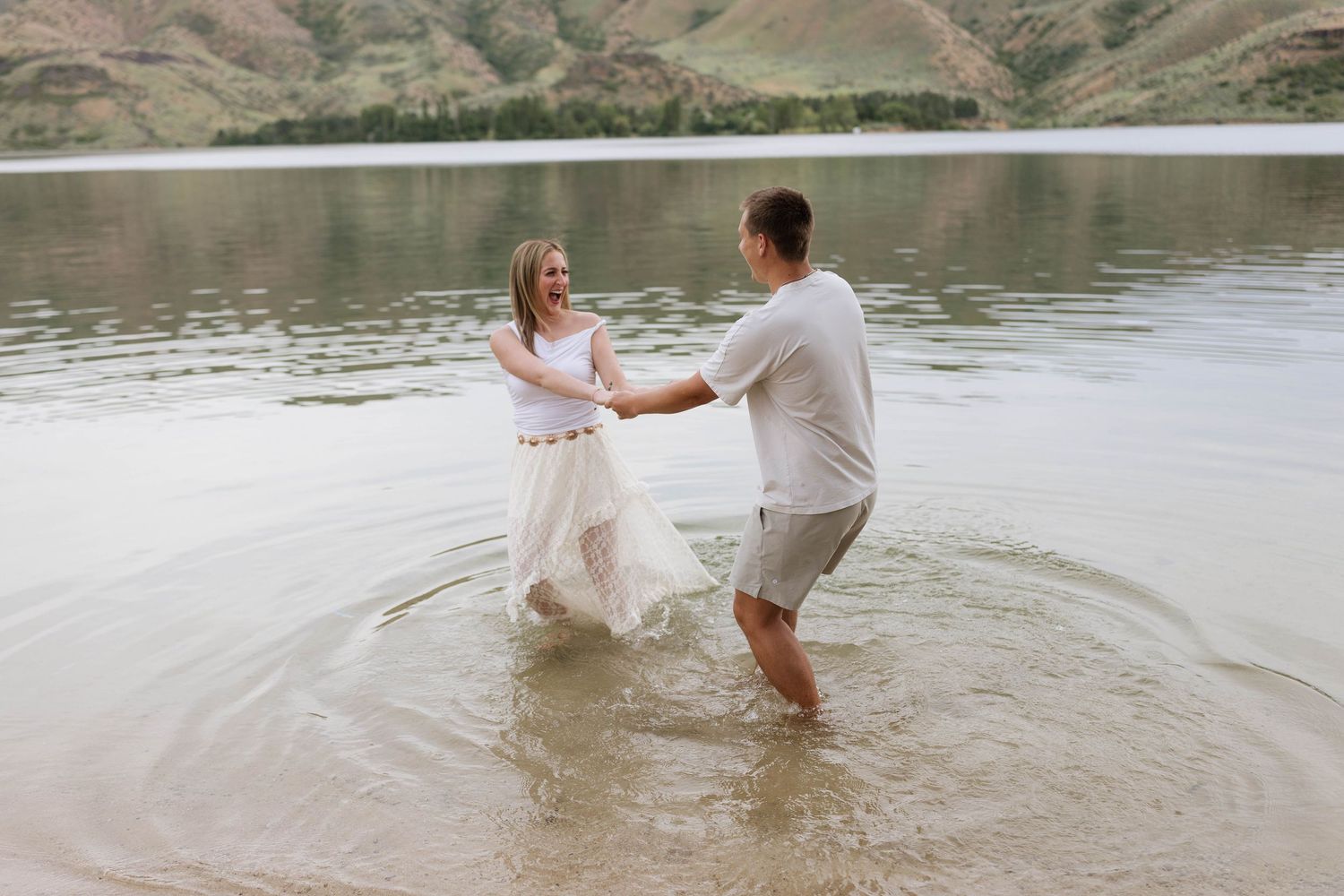 Couple dancing in shallow lake water, bride in white flowing dress, groom in casual gray outfit, scenic green hills backdrop.