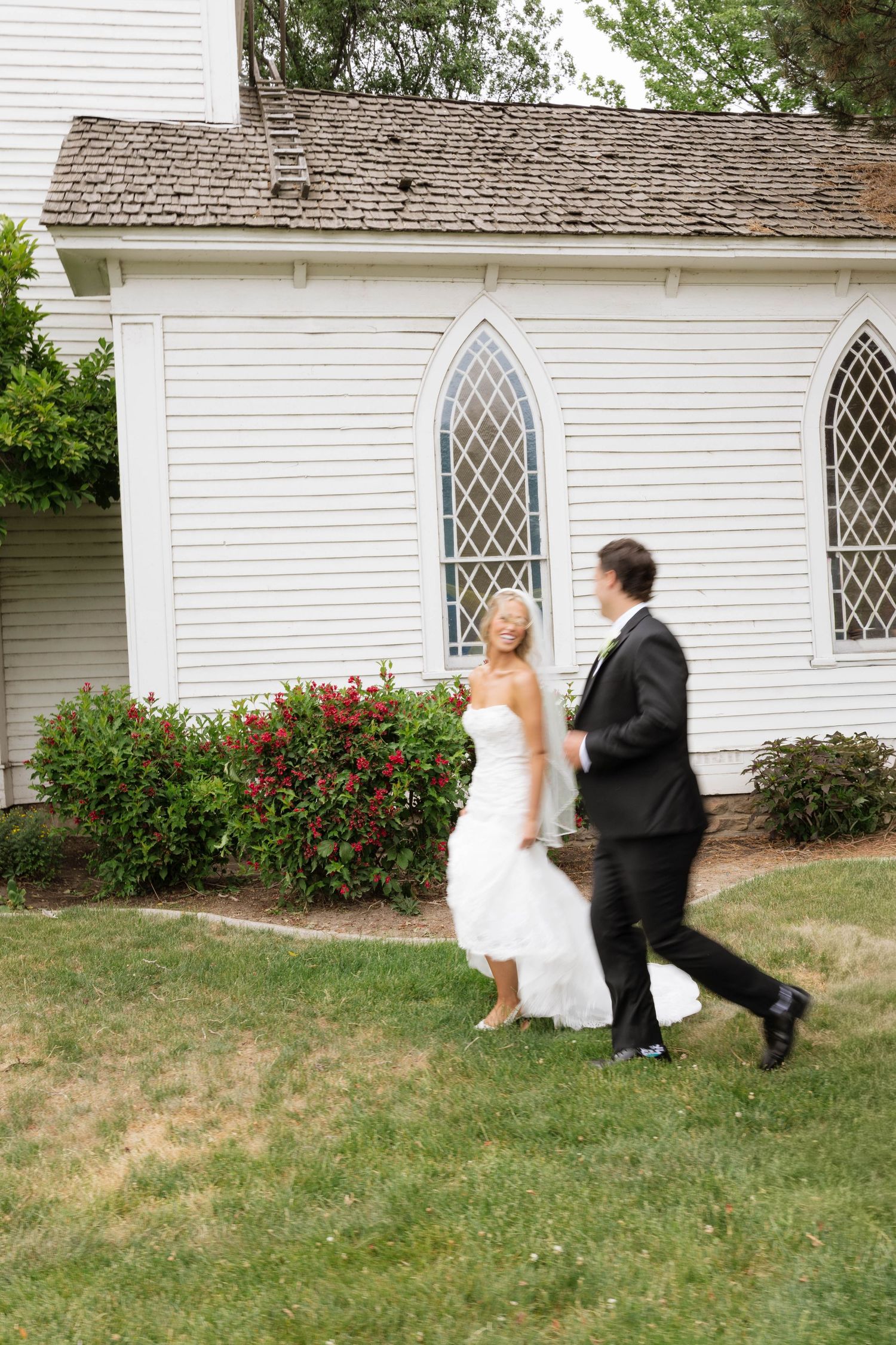 Bride and groom walking together on green grass outside a white chapel with gothic windows on their wedding day.