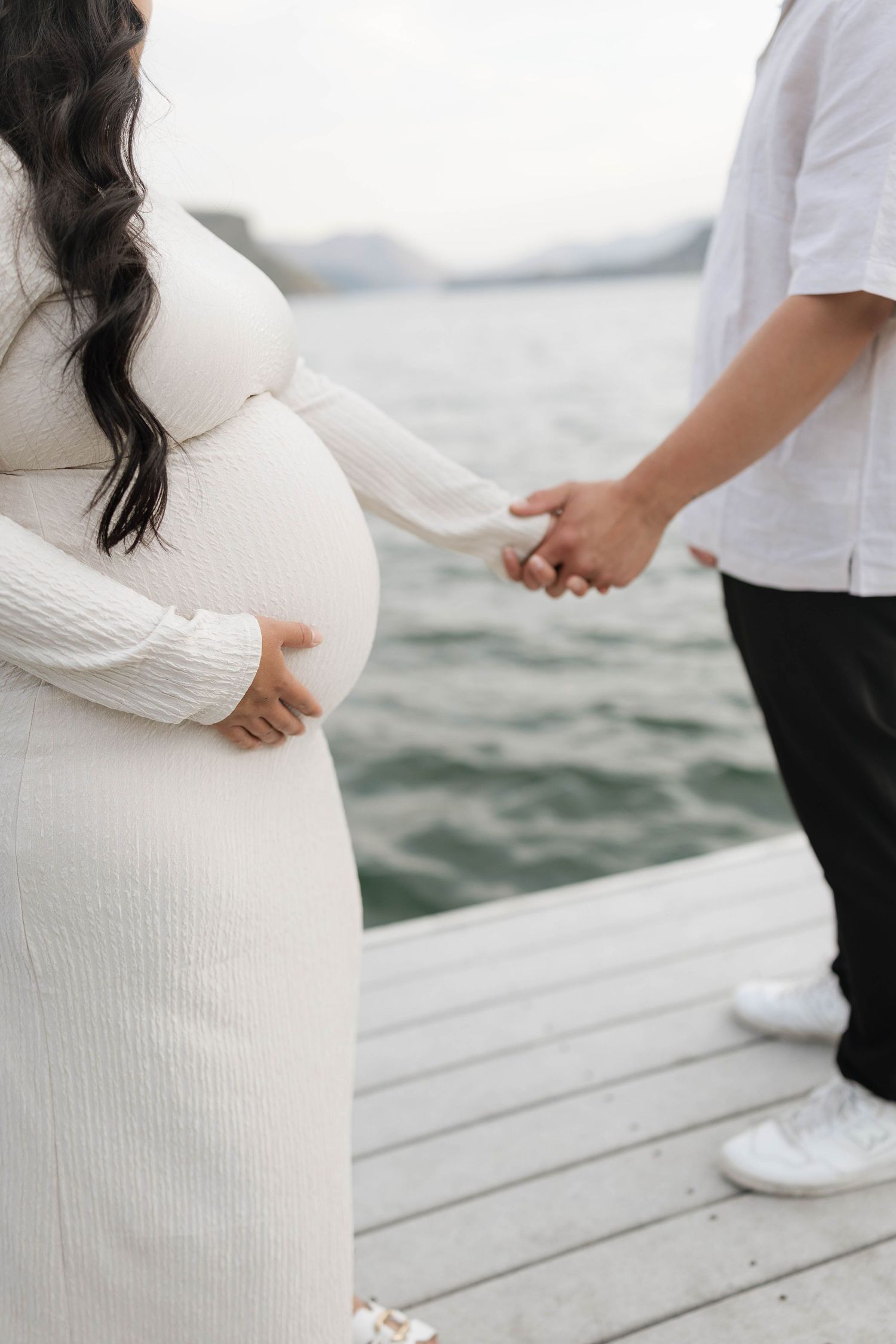 Pregnant woman in white dress holding belly with partner on waterfront dock during maternity photoshoot.