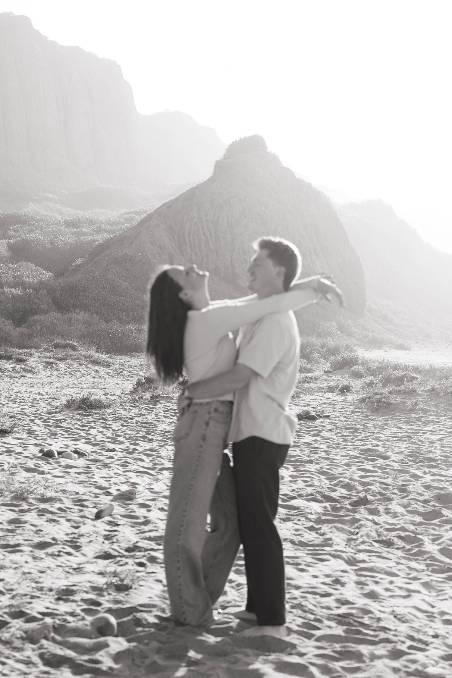A romantic couple embraces on a misty beach with dramatic cliffs in the background.