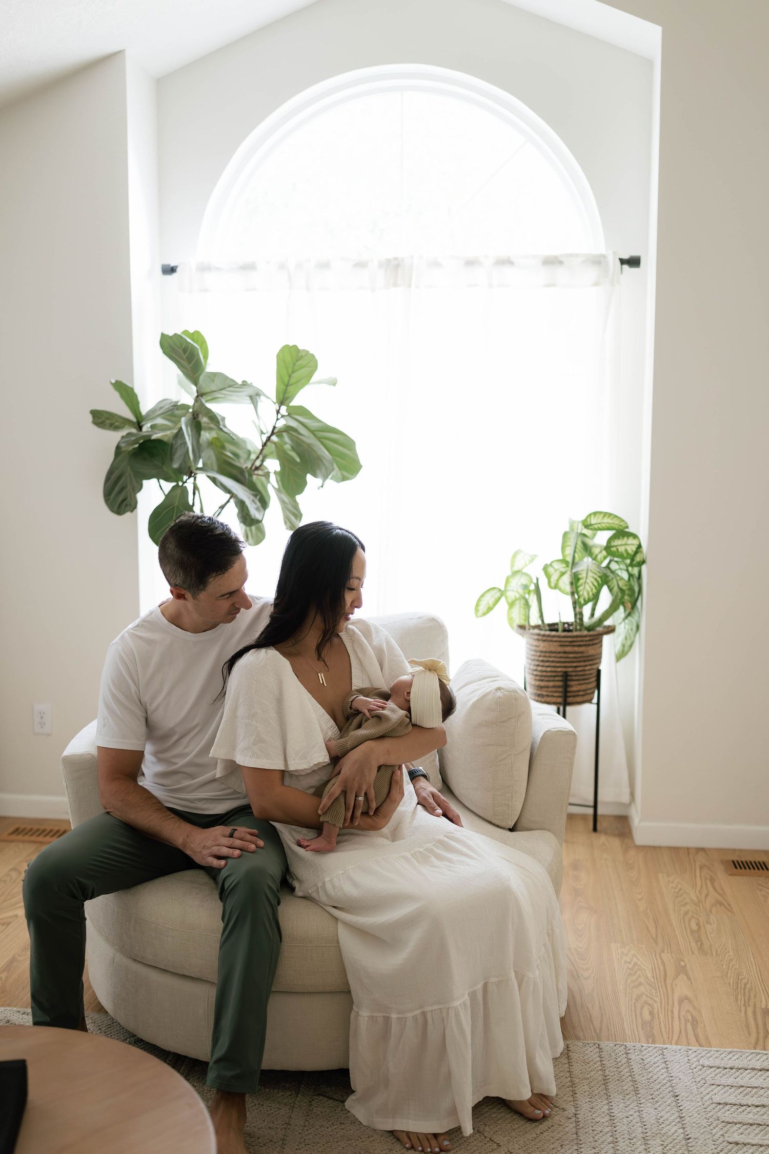 Parents sit on a cream sofa in a bright living room, mother nursing her newborn baby beside a large arched window and green plants.