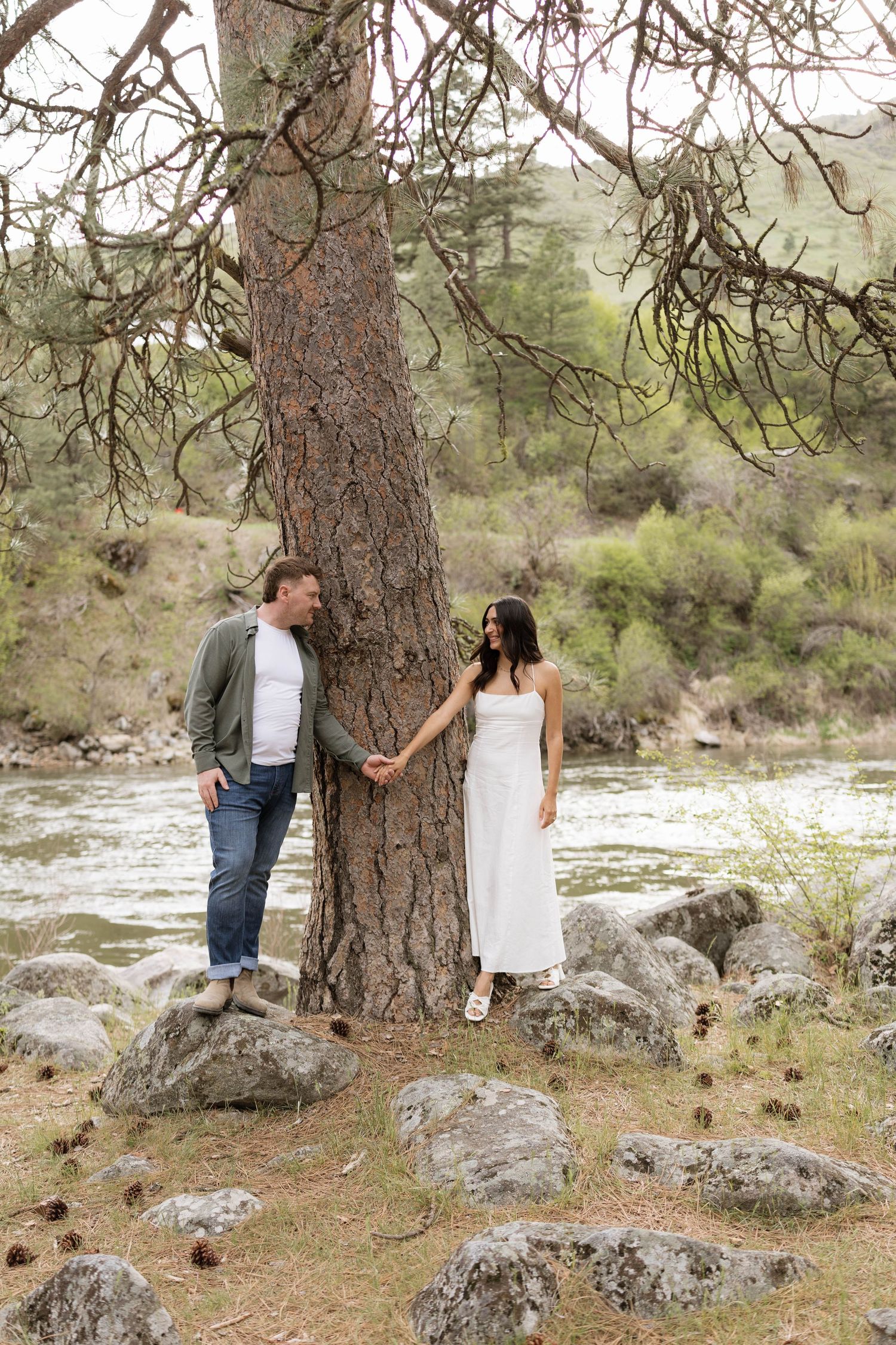Couple holding hands around a pine tree by a rocky riverbank, woman in white dress, man in green jacket and jeans.