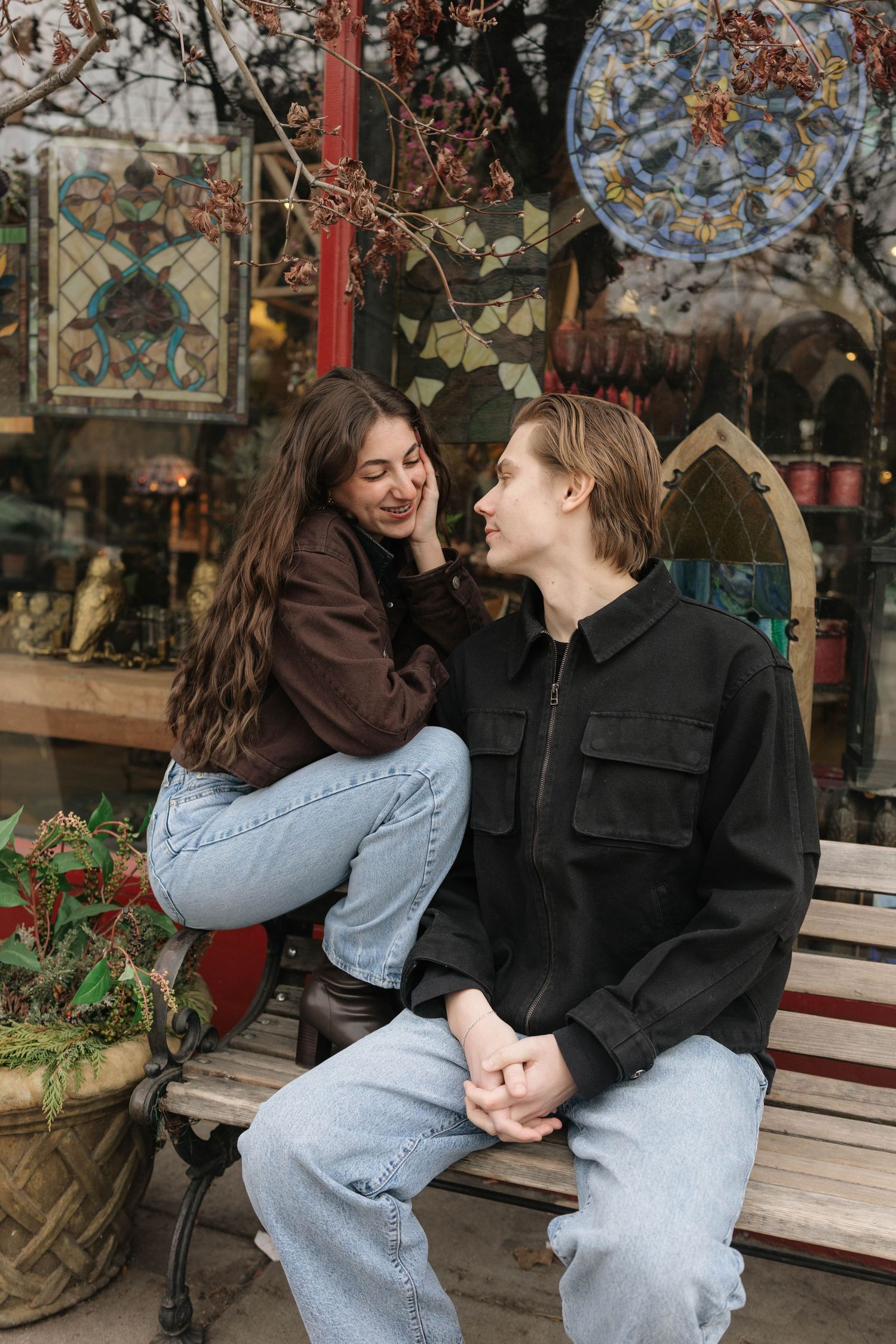 A couple in casual denim and dark jackets share intimate moments while sitting on a wooden bench outdoors.