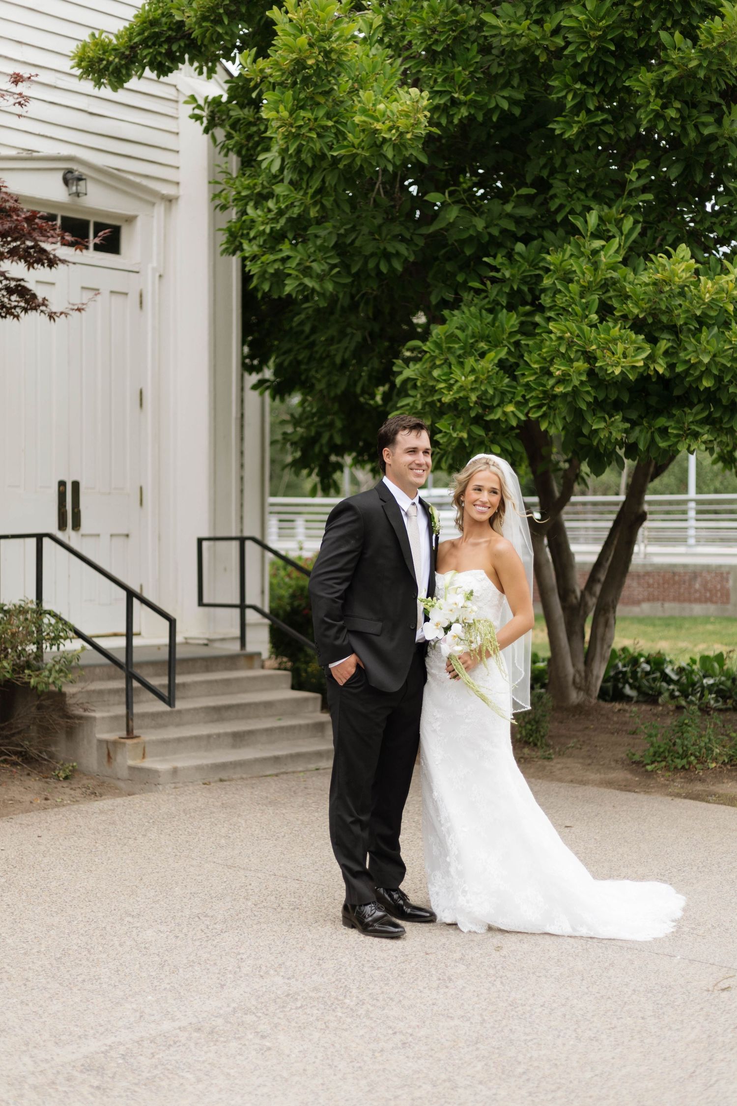 Bride and groom posing outside a white church, bride in strapless gown holding bouquet, groom in black tuxedo.