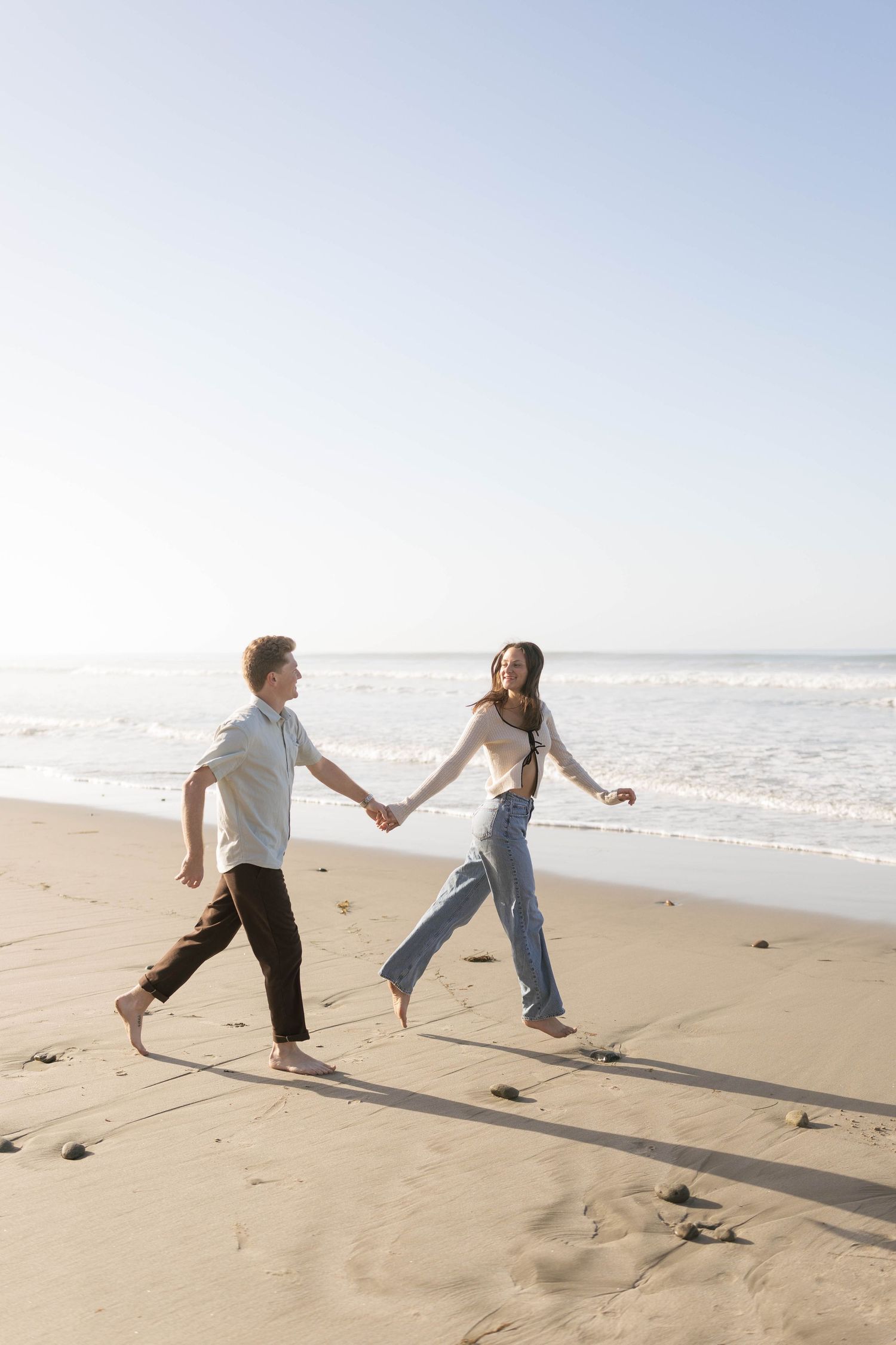 Two people playfully run and hold hands on a sunny beach with ocean waves in the background.