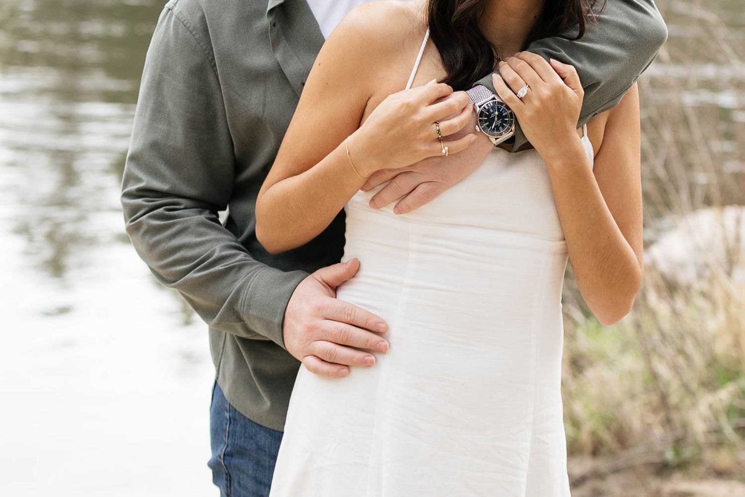 Couple embracing outdoors near water, woman in white dress with man in green shirt wearing a watch.