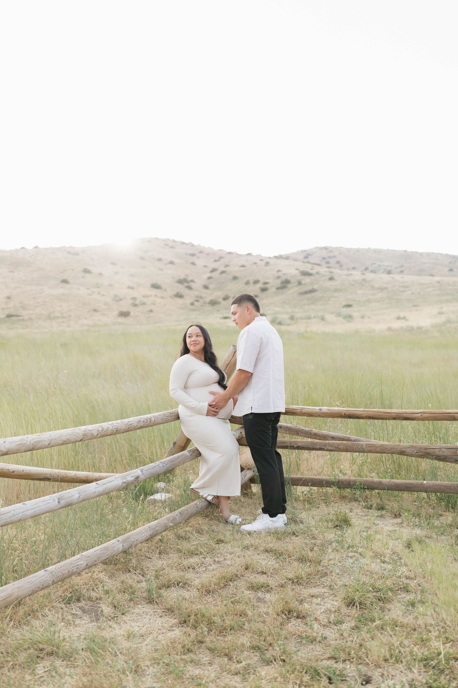Pregnant couple poses by rustic fence in grassy field, woman in white dress cradling baby bump, golden hour light.