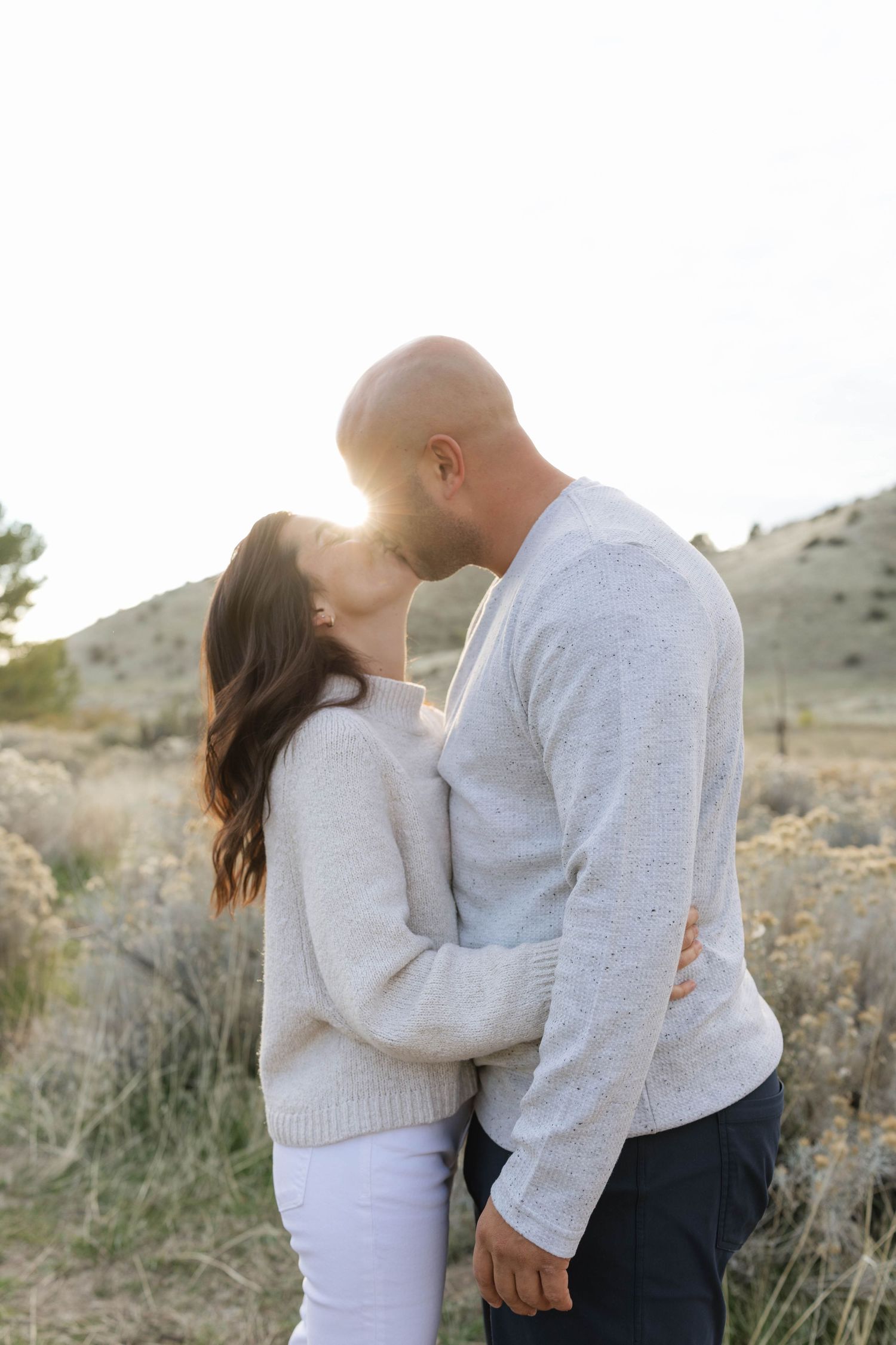 A romantic couple in matching light sweaters embrace in a desert landscape at sunset.