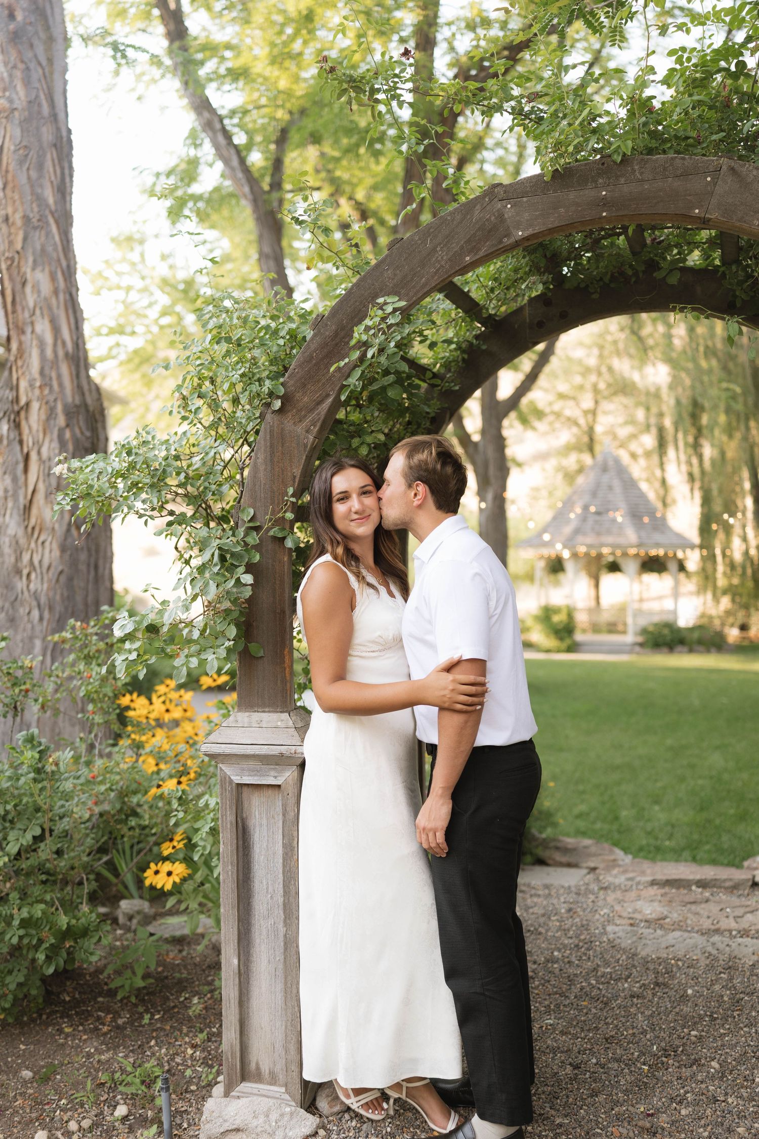 Couple embracing under a wooden arch in a lush garden, bride in white dress, groom in white shirt and black pants.