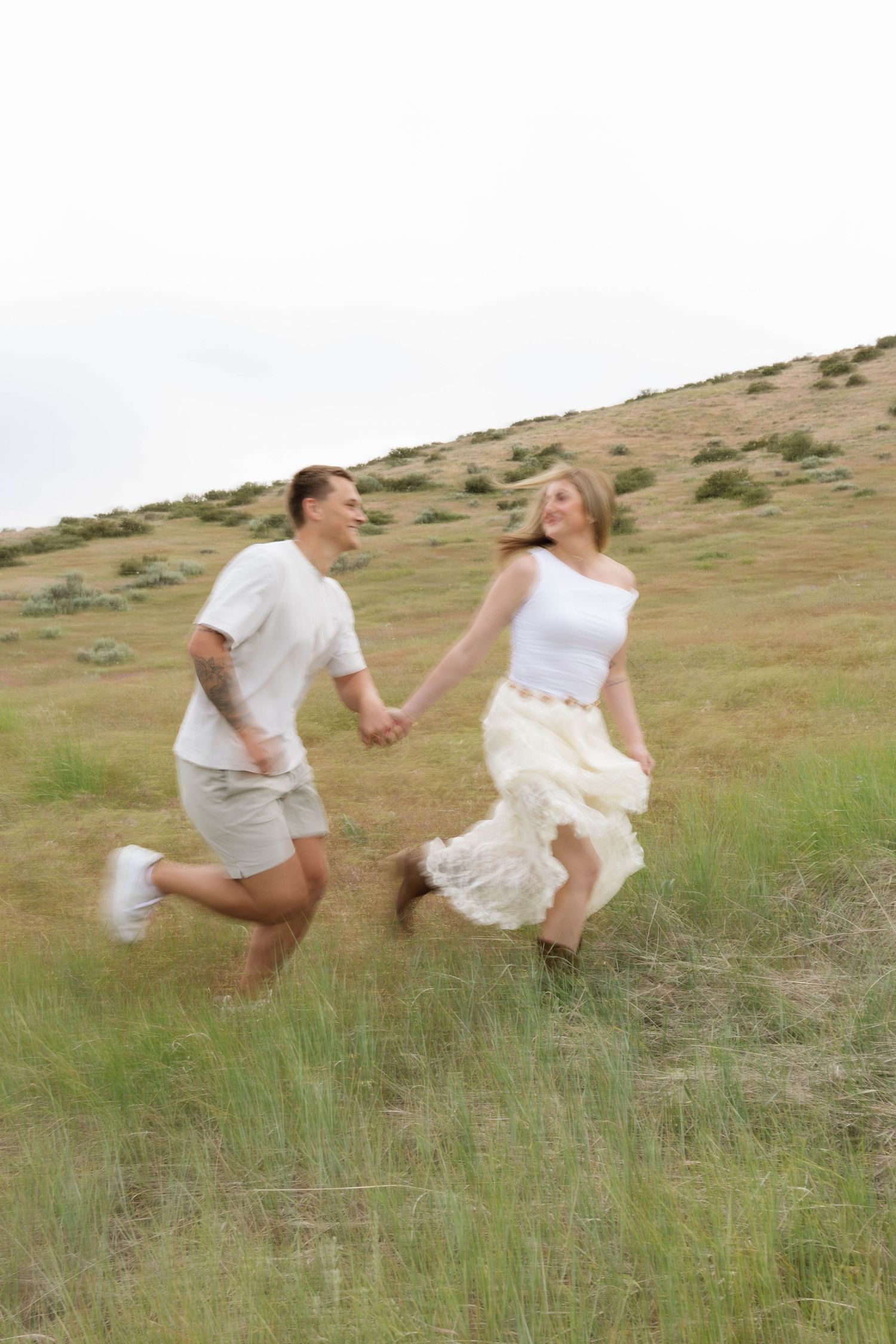 A joyful couple runs hand-in-hand through a grassy hillside, wearing casual white outfits on an overcast day.
