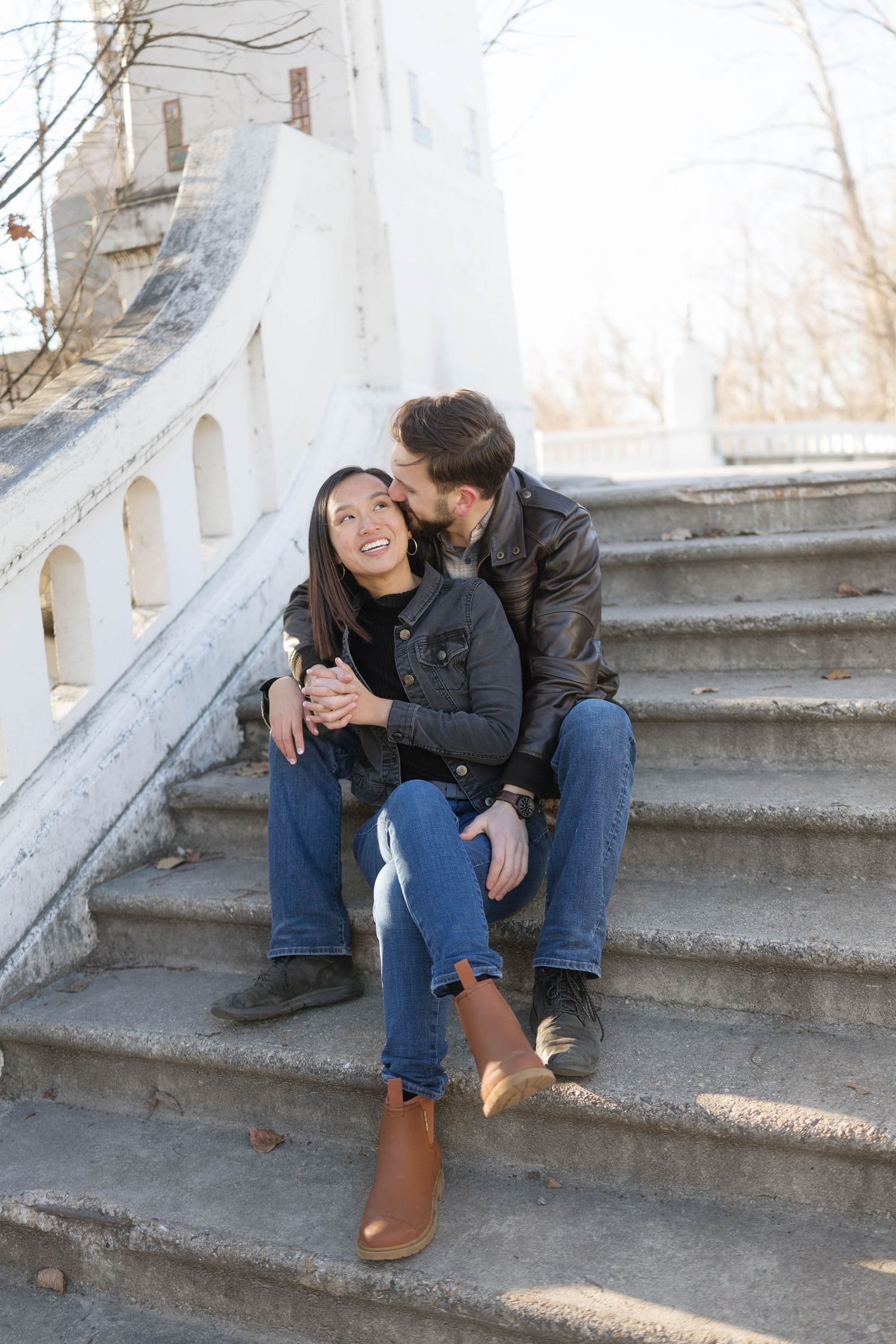 A couple sits together on concrete steps during a sunny autumn day, sharing a tender moment.