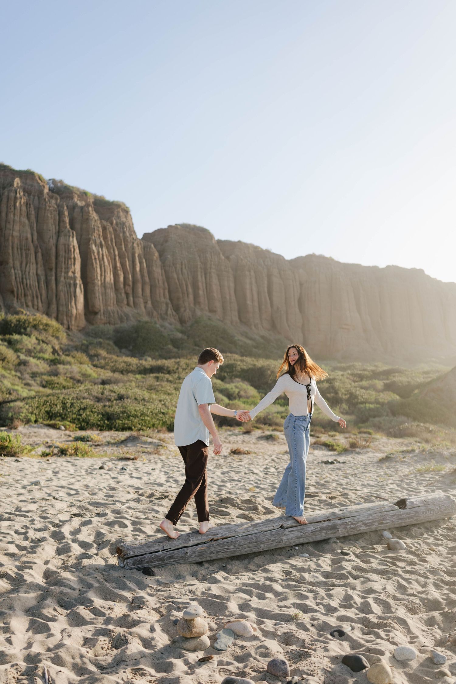 A couple walks together on a fallen log at a scenic desert beach with towering cliffs in the background.