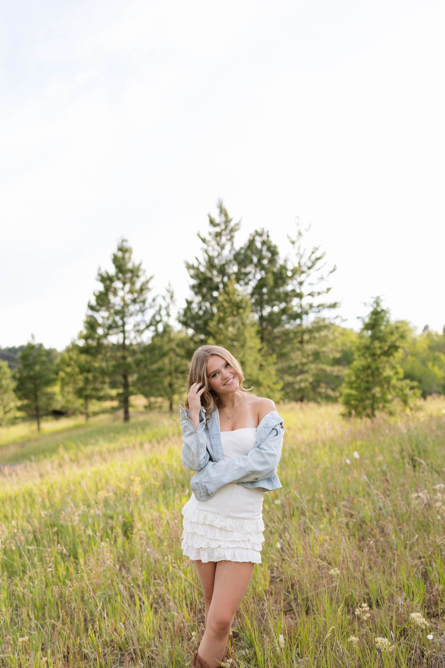 Young blonde woman in white dress and blue jacket smiling in a sunny meadow with pine trees in background.