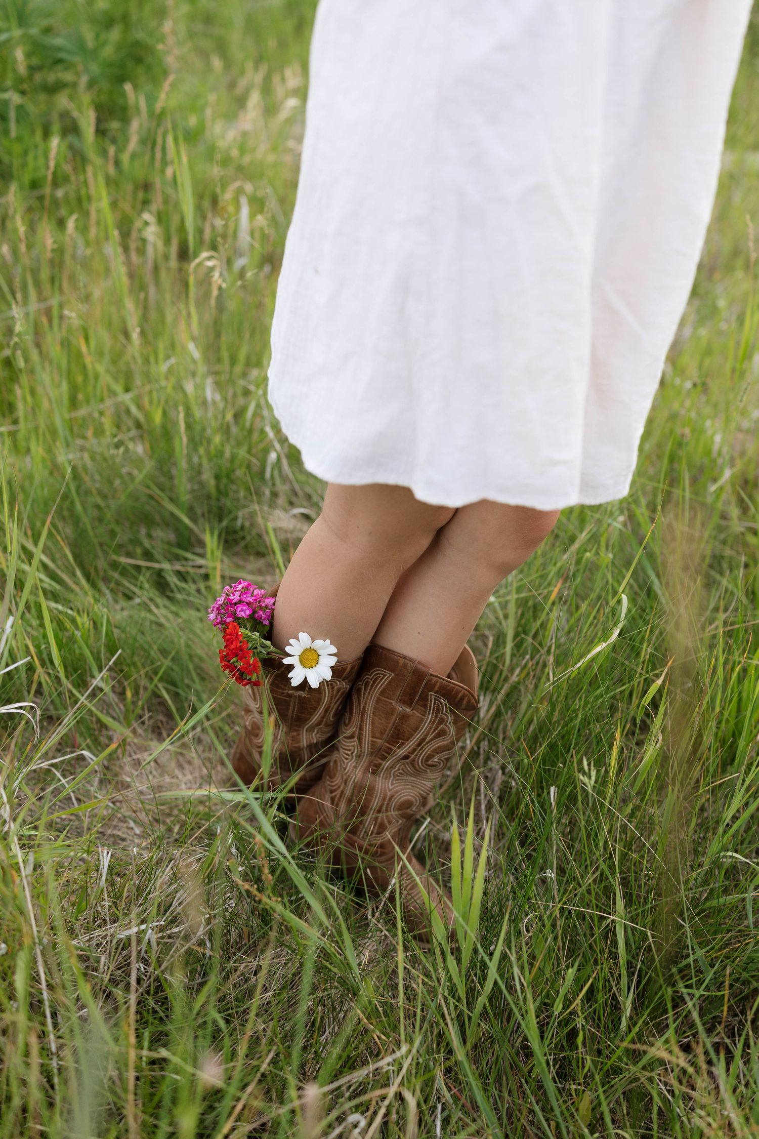 Person wearing brown cowboy boots with wildflowers tucked inside, standing in green grass field wearing a white dress.