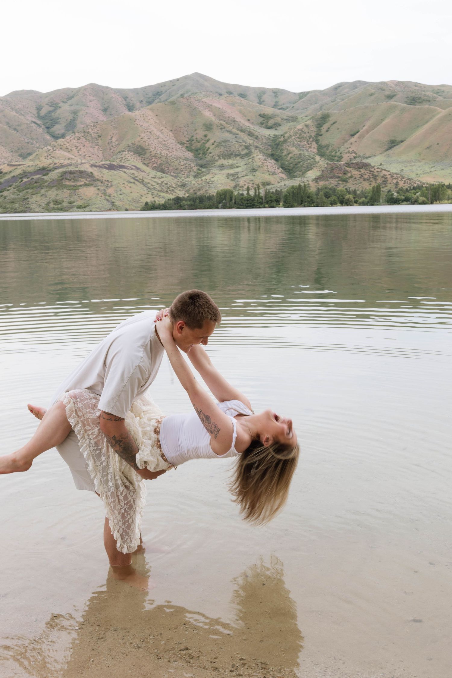 Couple dancing romantically in shallow lake water with scenic mountain backdrop on a cloudy day.