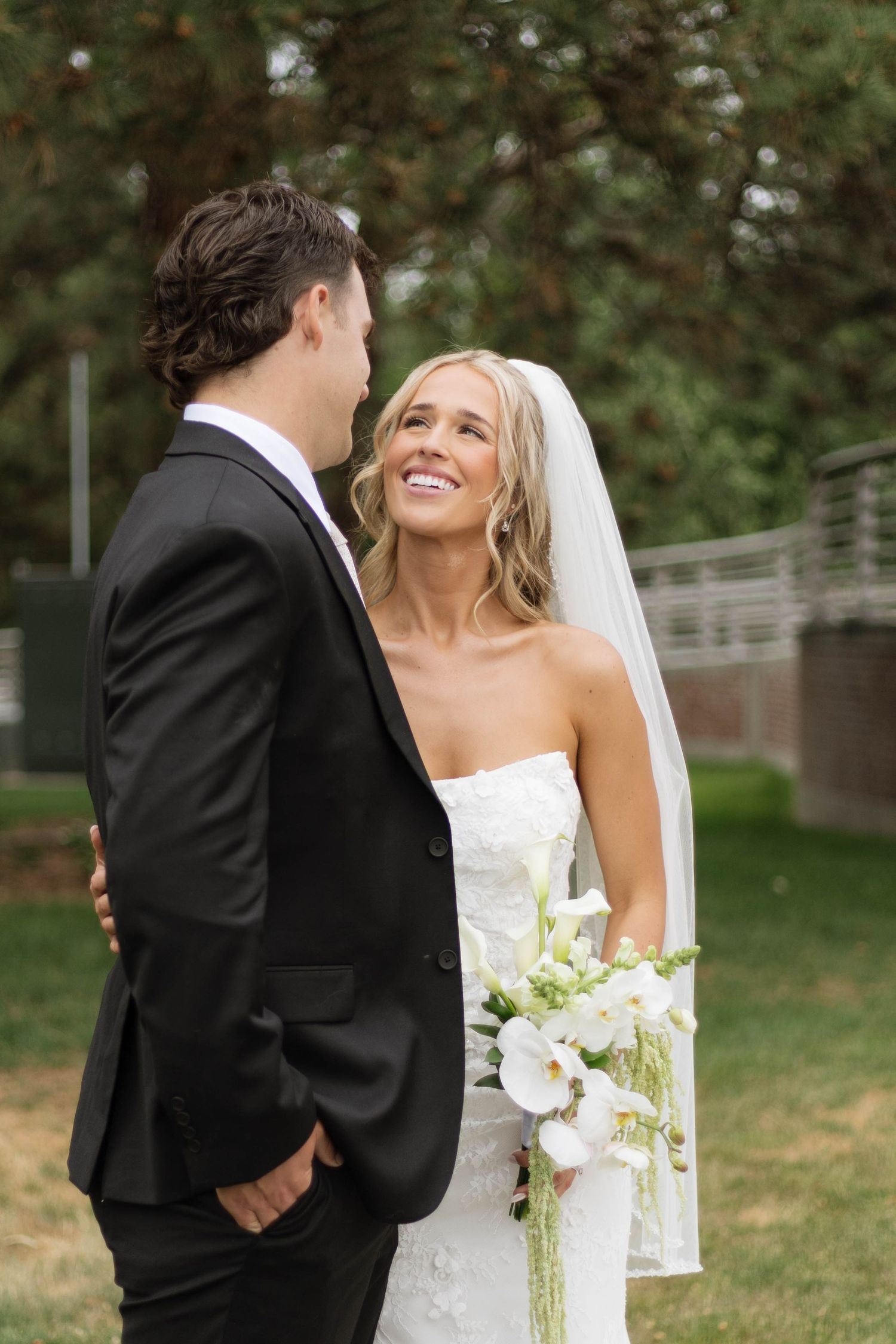 Smiling bride in white strapless gown and veil holds bouquet while gazing at groom in black suit outdoors.