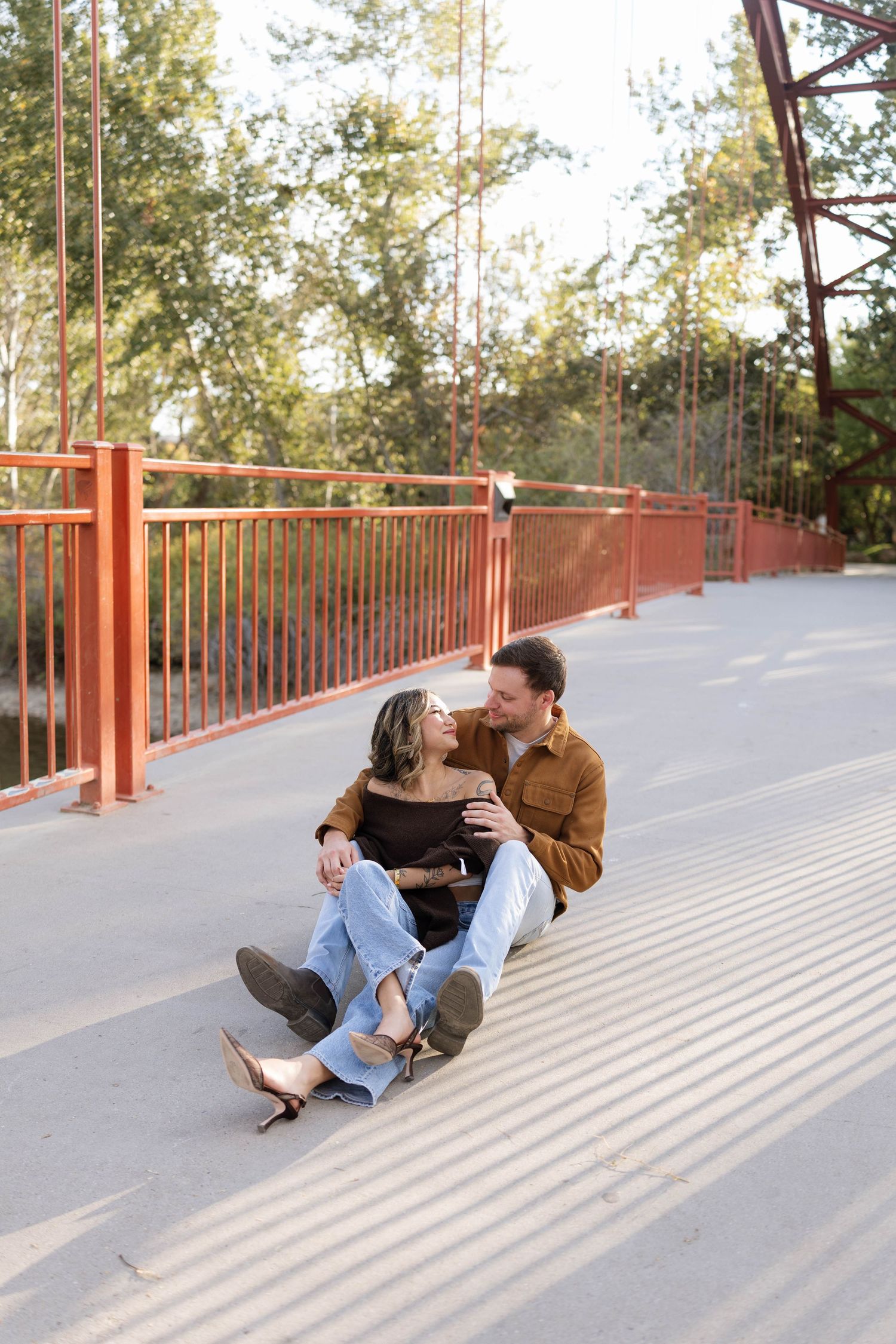 Couple sitting close and embracing on a sunlit bridge deck with rust-red railings and green trees in the background.
