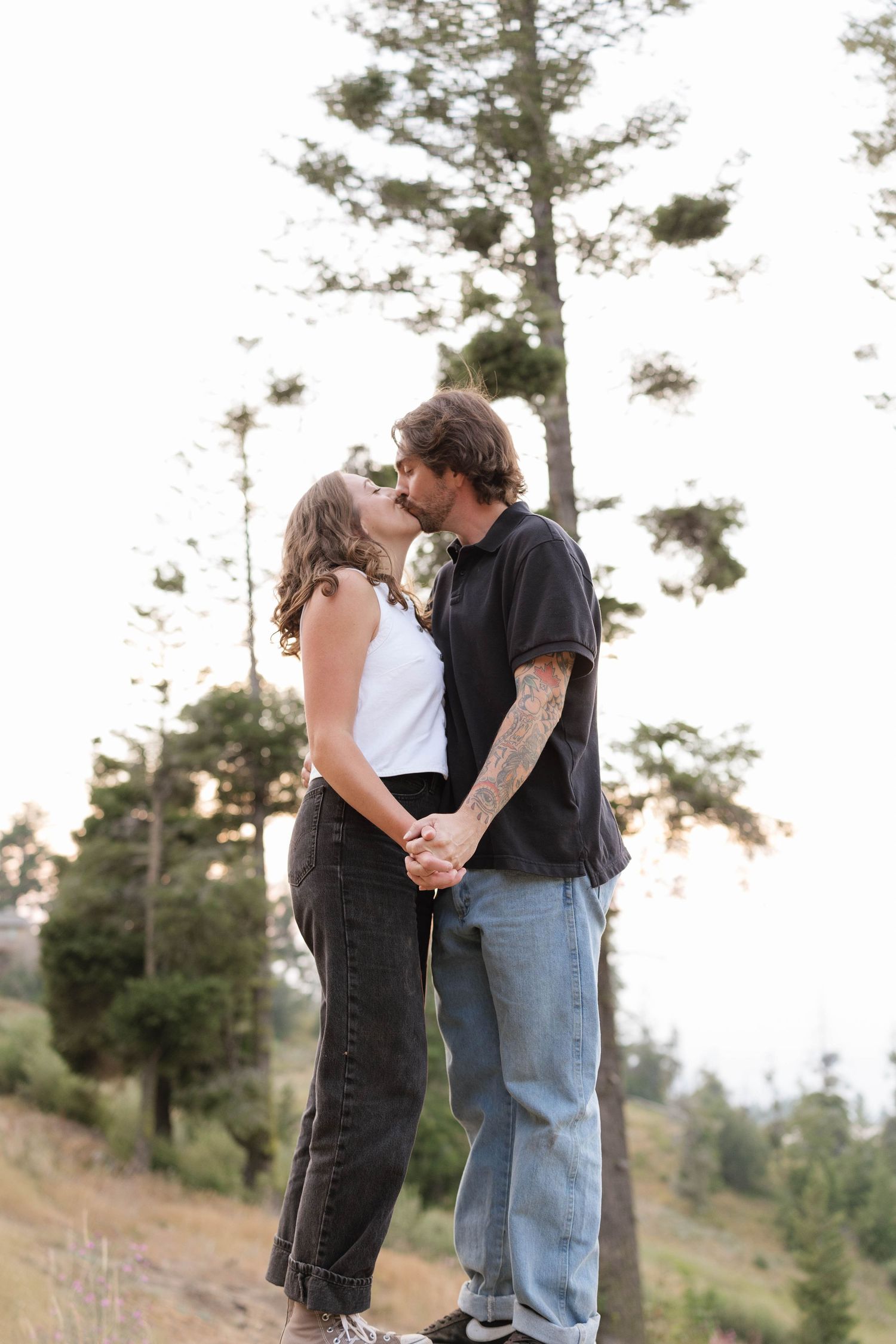 A couple shares a kiss while holding hands outdoors, standing among tall pine trees on a grassy hillside at sunset.