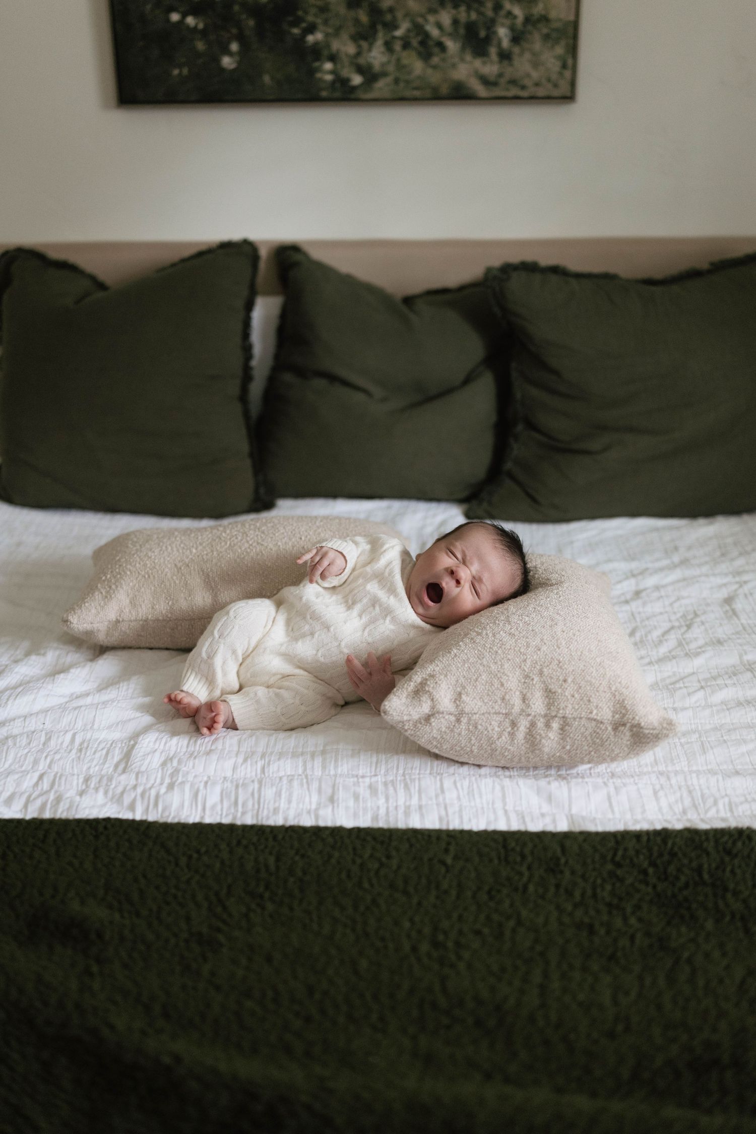 A baby yawning while lying on a white bed with olive green pillows and blanket.