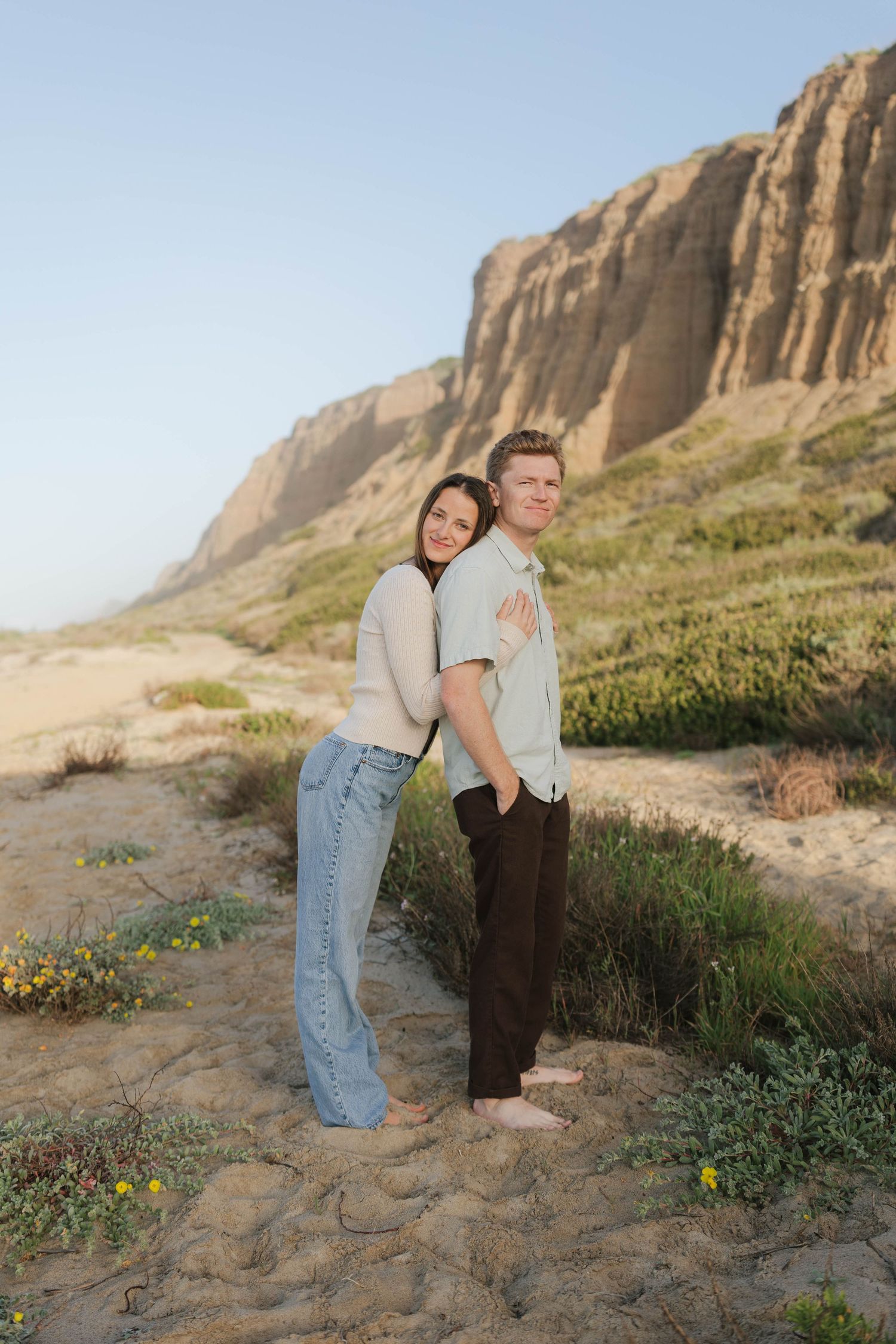 A couple stands together on a sandy beach path with dramatic coastal cliffs and wildflowers in the background.