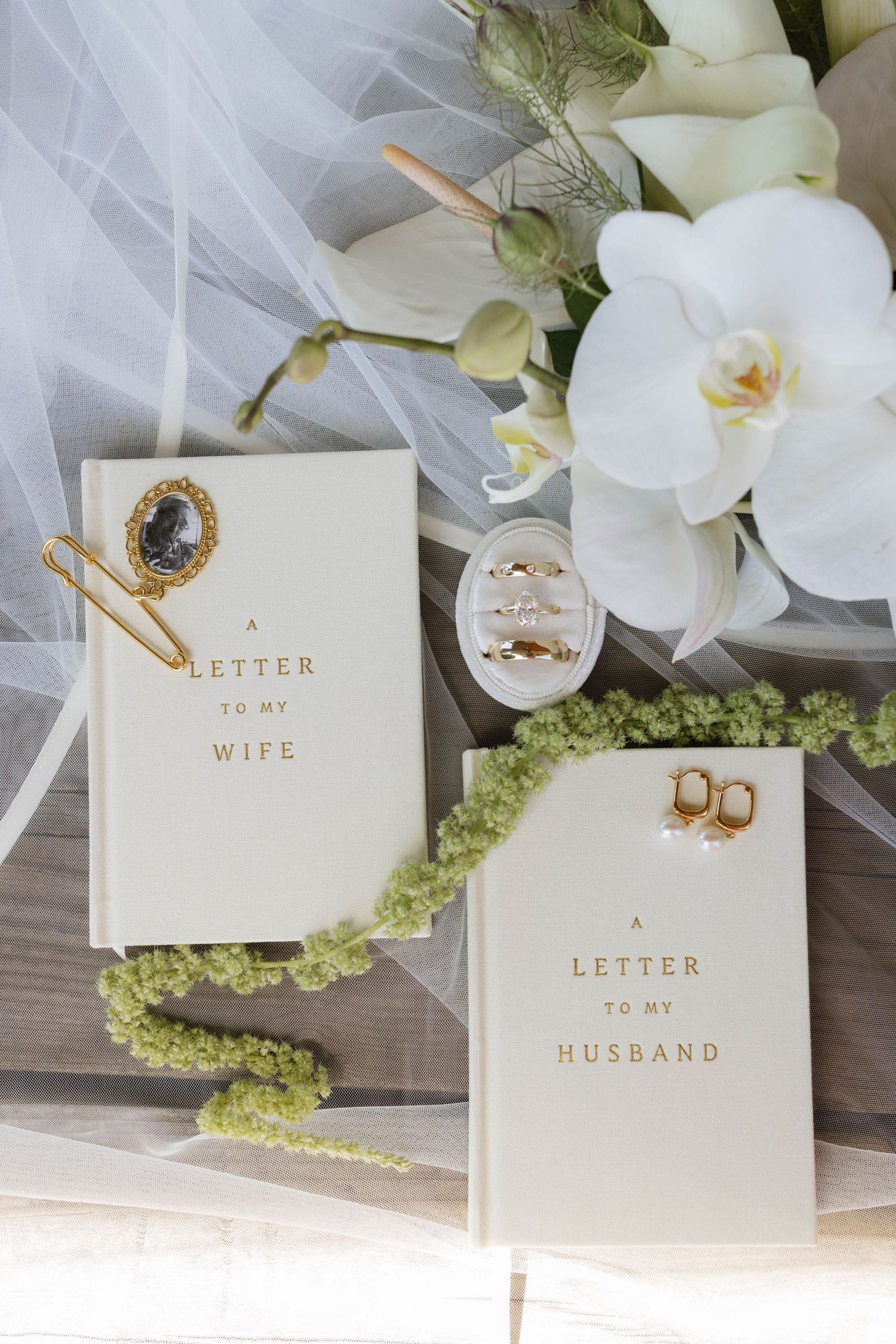 Wedding flat lay with letter books to wife and husband, gold rings, earrings, white orchids, and bridal veil.