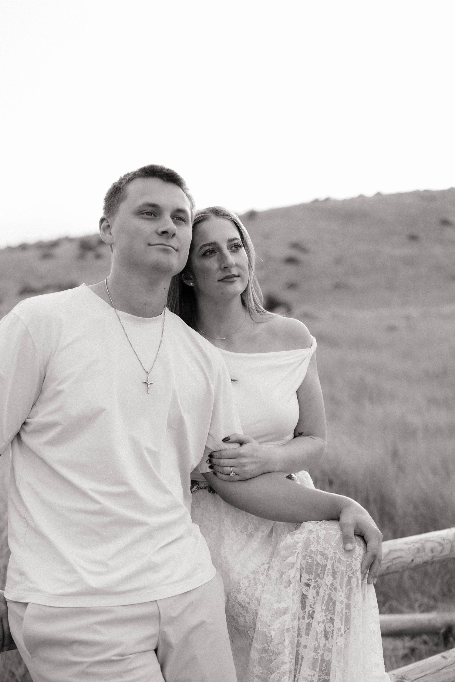 Black and white portrait of a young couple in white outfits leaning on a wooden fence with hills behind them.