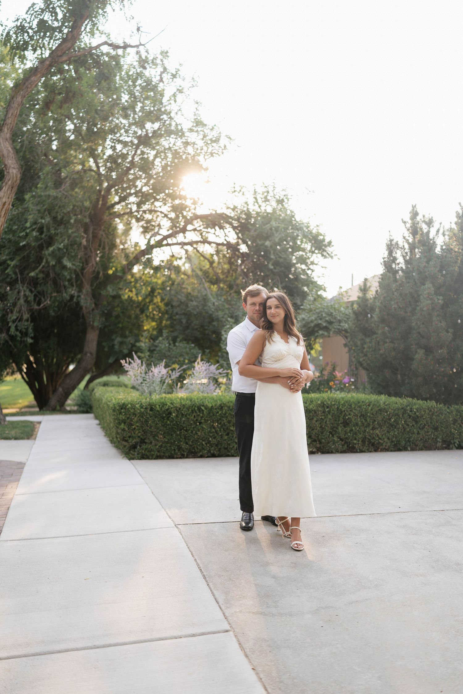 Couple embracing at sunset in a lush garden, woman in white dress and man in white shirt and black pants.