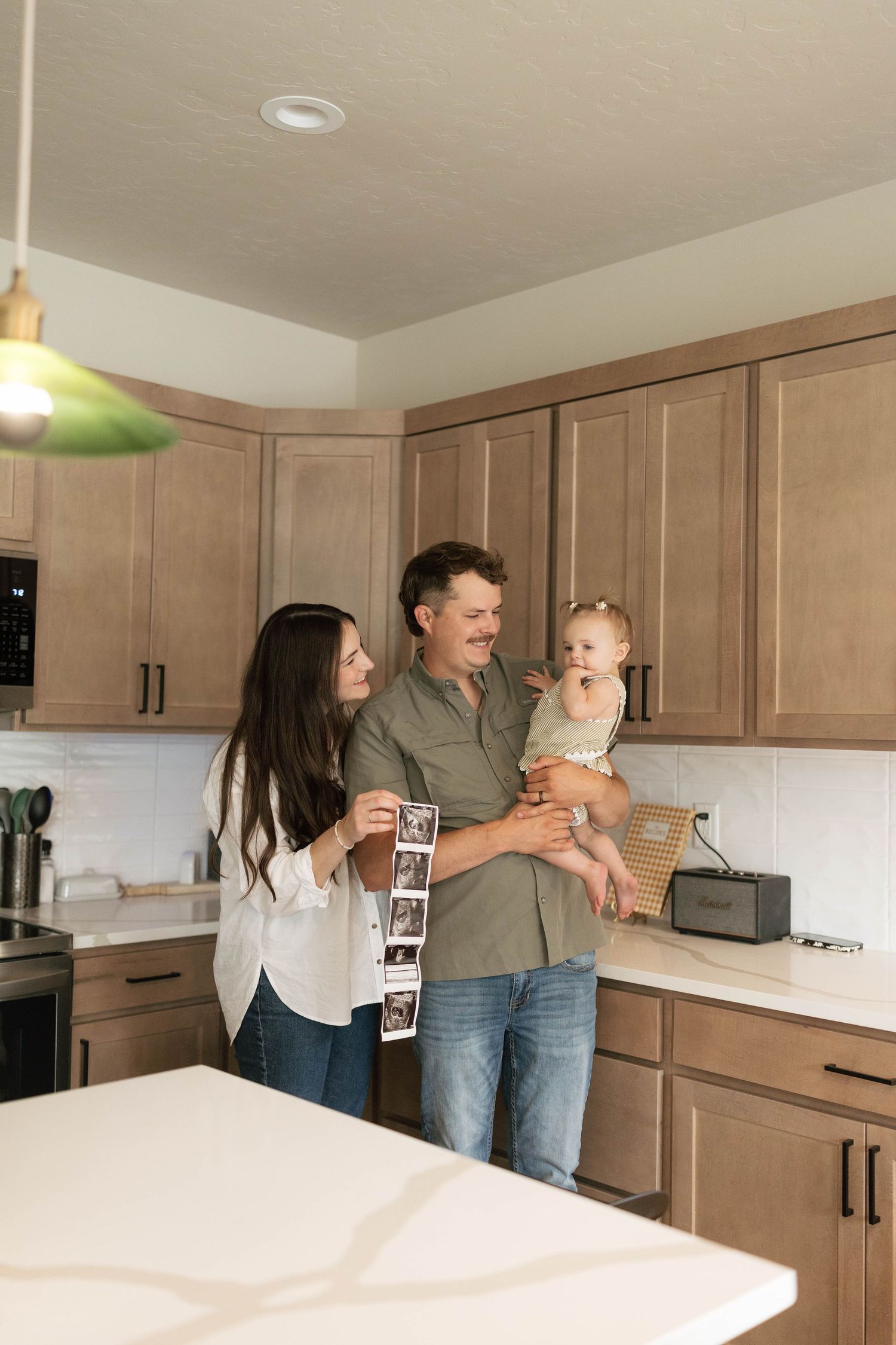 A happy couple holding their baby and ultrasound photos in a modern kitchen, announcing a new pregnancy.