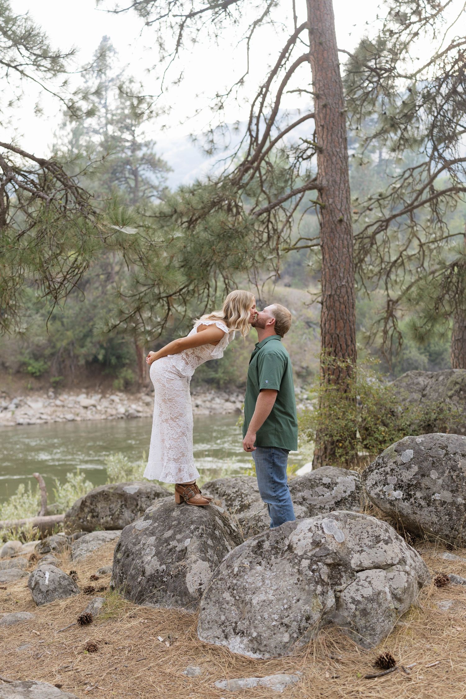 Couple sharing a romantic kiss on rocks near a river, surrounded by pine trees in a serene forest setting.