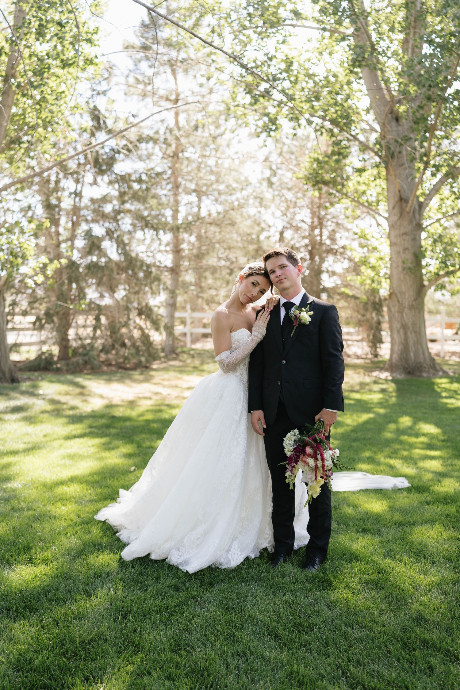 Bride in white lace gown leans on groom in black suit holding bouquet, posing outdoors on green grass with trees.