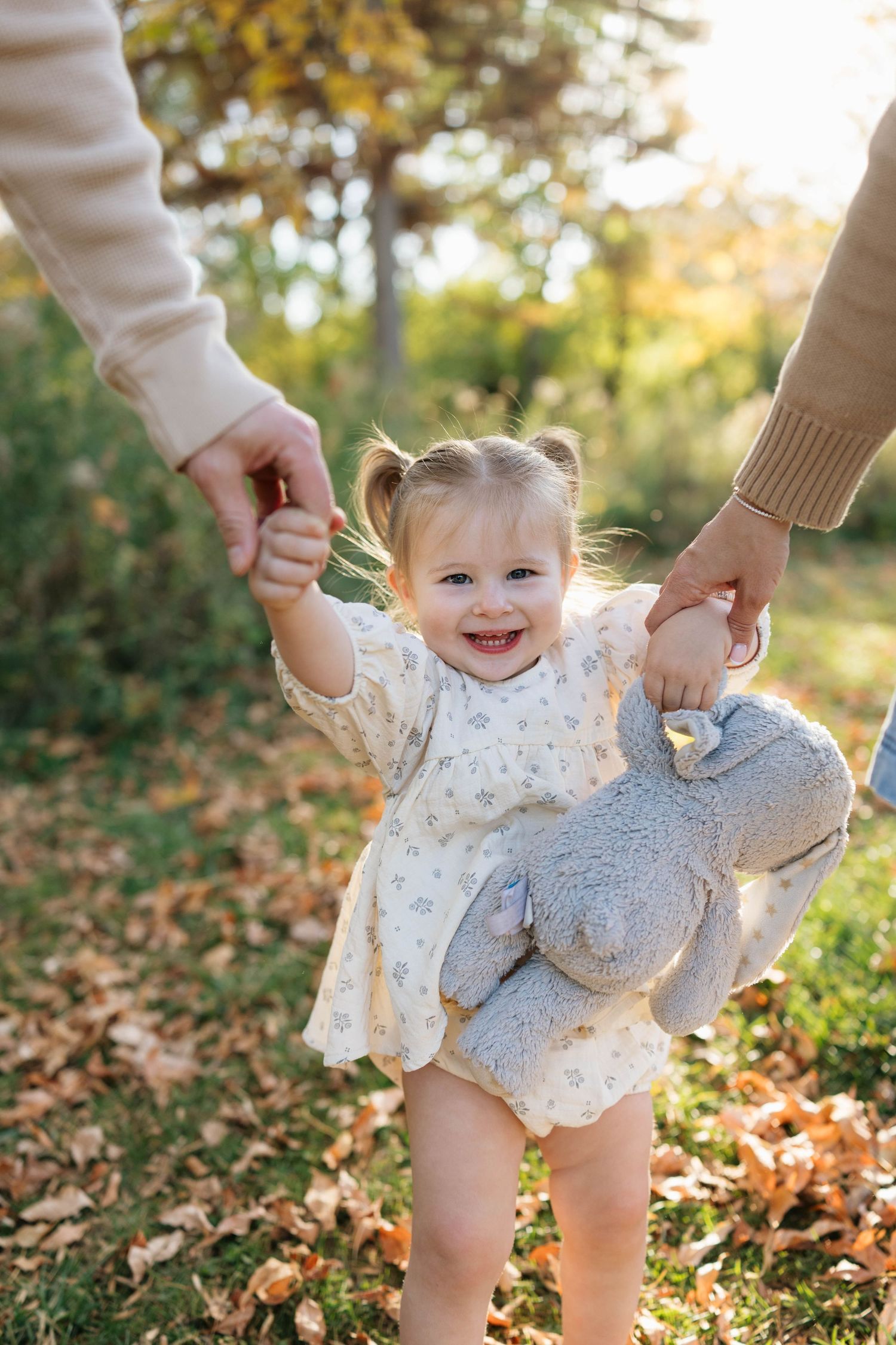 Smiling toddler girl in a floral dress holds a gray stuffed bunny while walking hand in hand with two adults on autumn leaves.