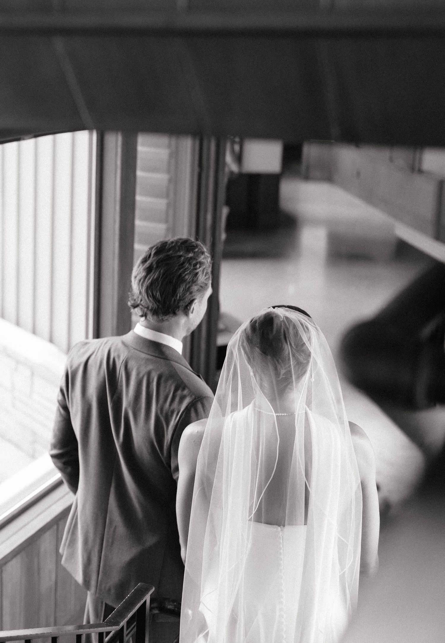 Black and white photo of a bride in veil and groom in suit seen from behind at their wedding venue.