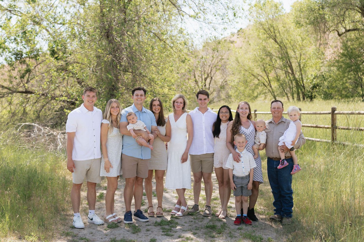 Large extended family group portrait on a dirt path surrounded by lush green trees and a wooden fence.