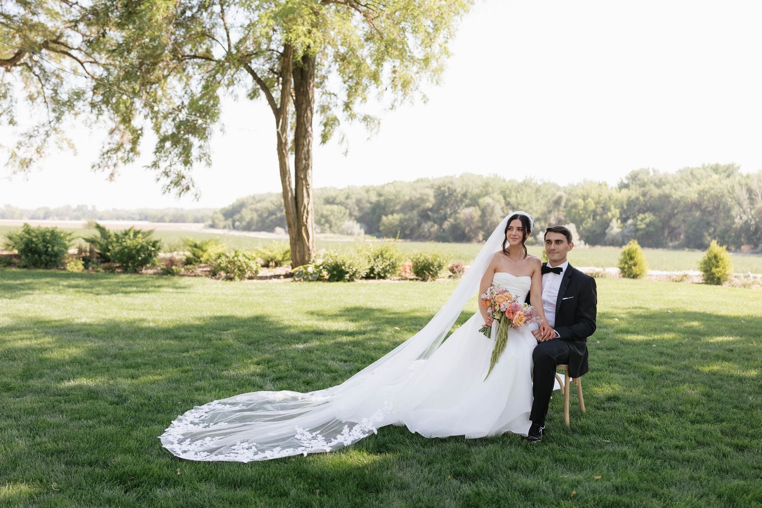 Bride in white gown with long veil sits beside groom in tuxedo on green lawn under a large tree.
